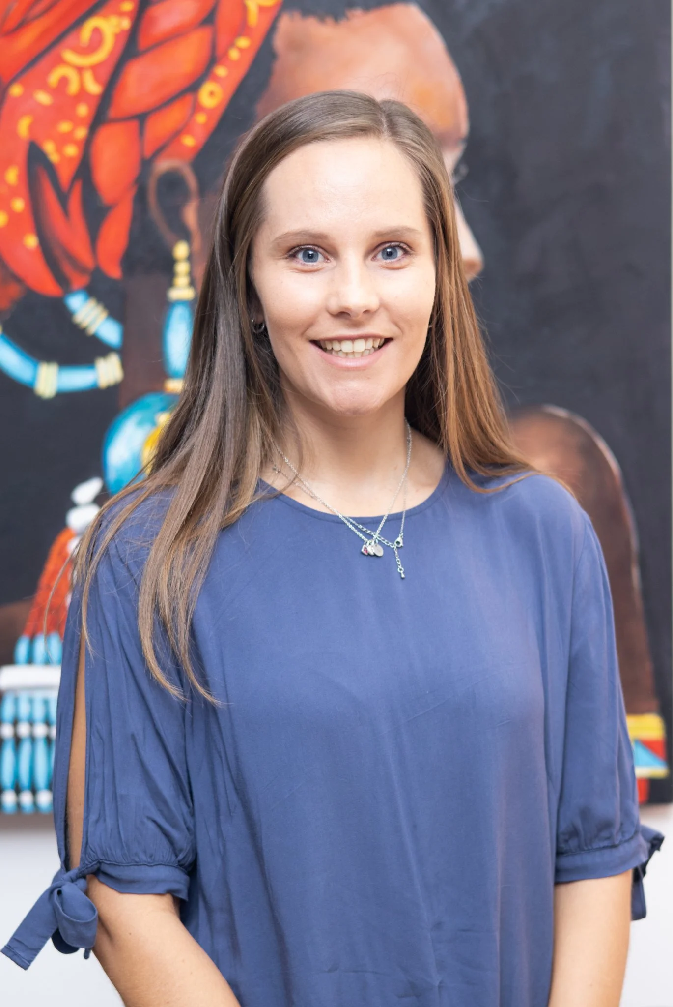 Young woman with light brown hair, blue eyes, wearing a blue blouse with tied sleeves and layered necklaces, smiling in front of a black background with colorful artwork.