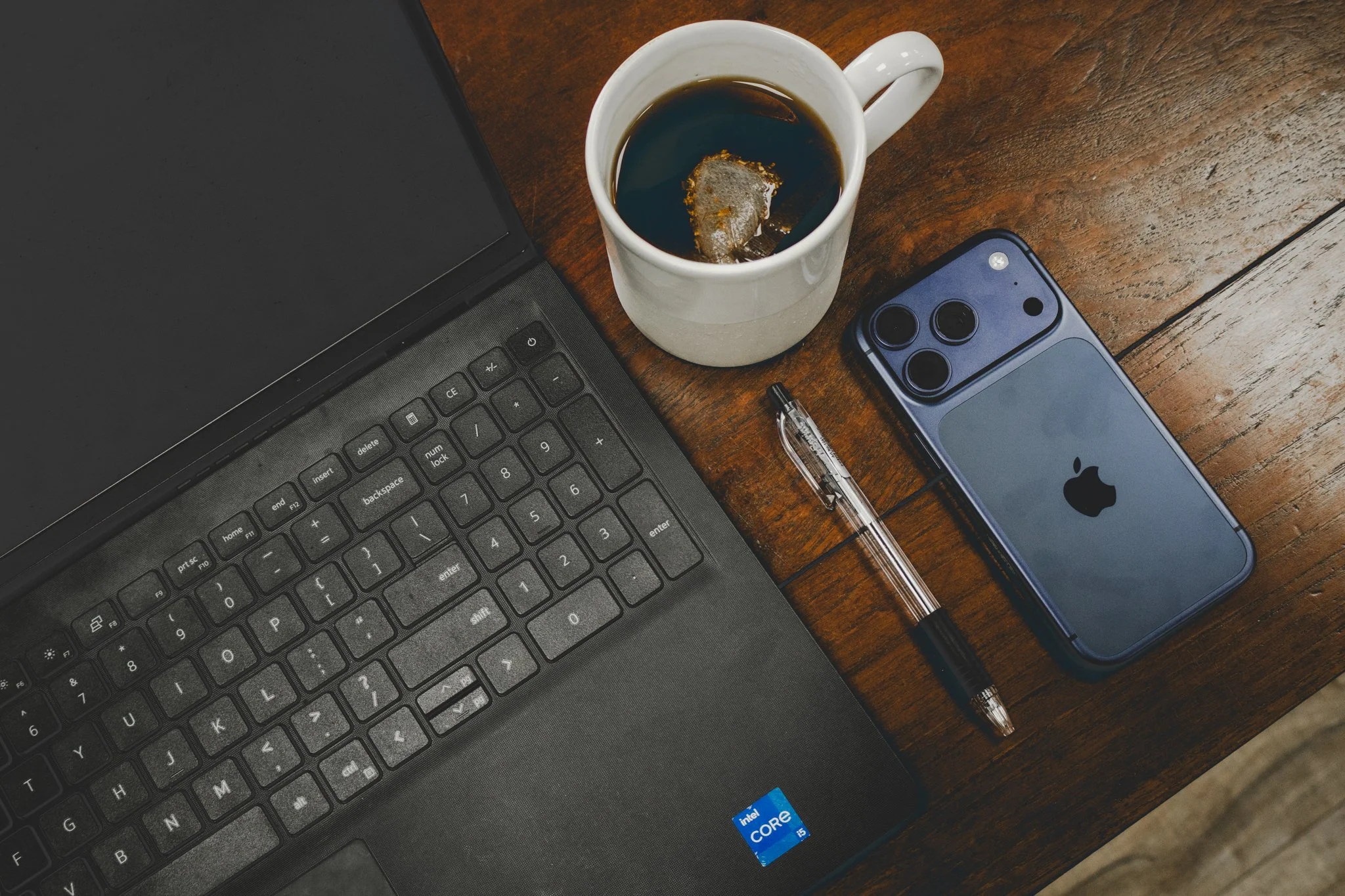Laptop, coffee mug with tea bag, black pen, iPhone with triple camera, on a wooden desk
