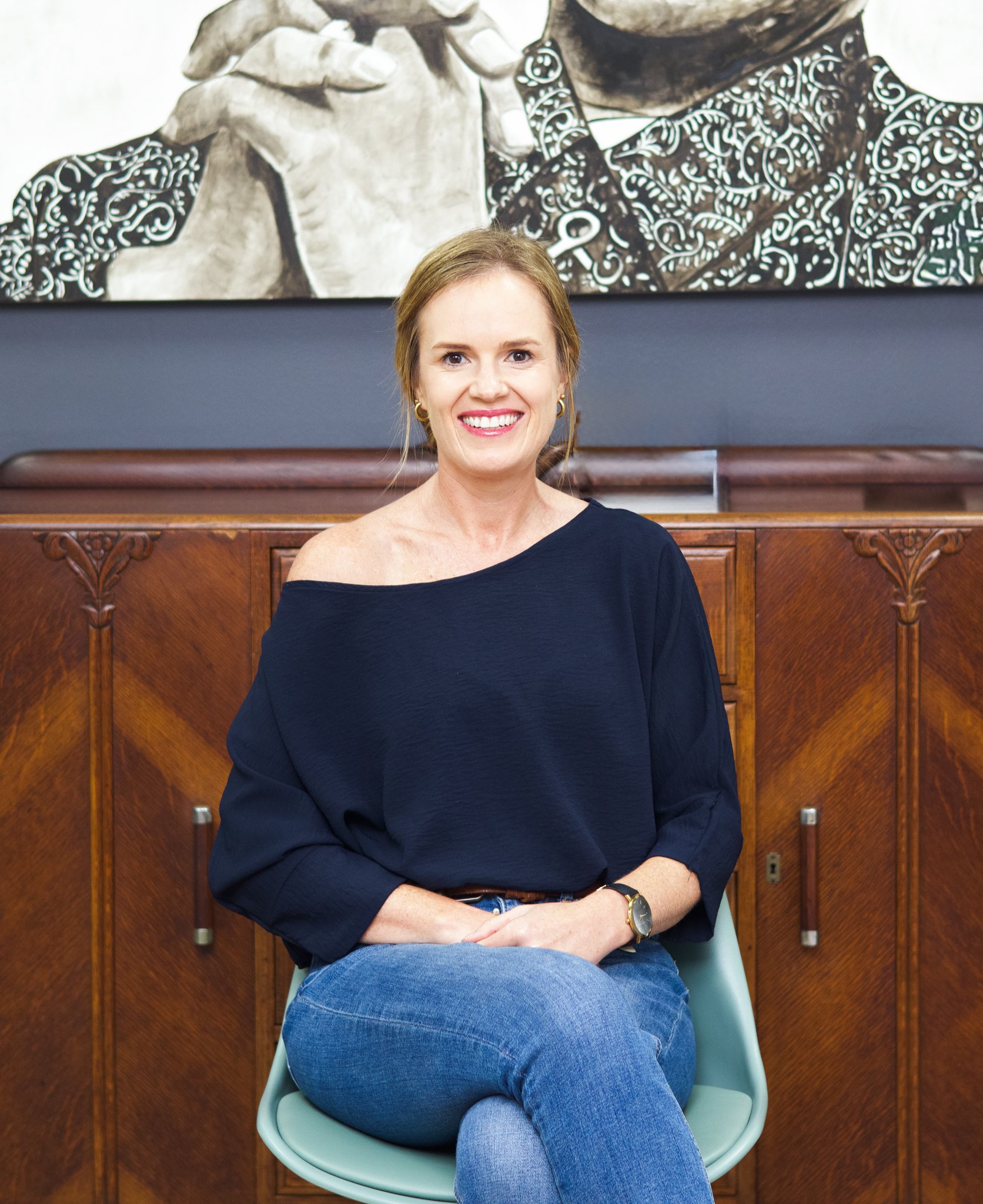 A smiling woman with blonde hair sitting on a light green chair, wearing a black off-shoulder top and blue jeans, with a wooden cabinet and a black and white portrait painting behind her.