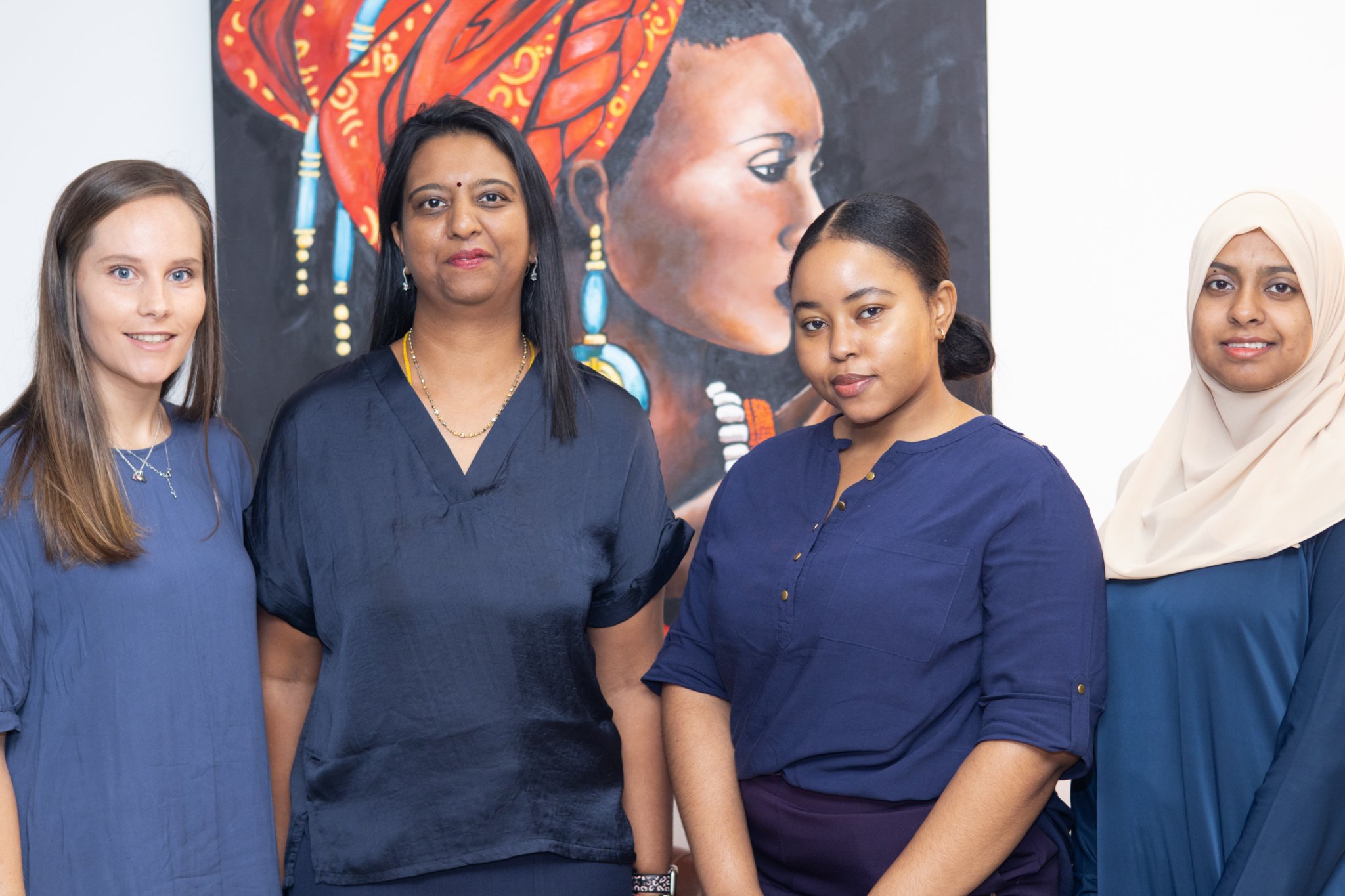 Four women standing together in front of a black artwork depicting a profile of a woman with colorful decorated headwrap and jewelry.
