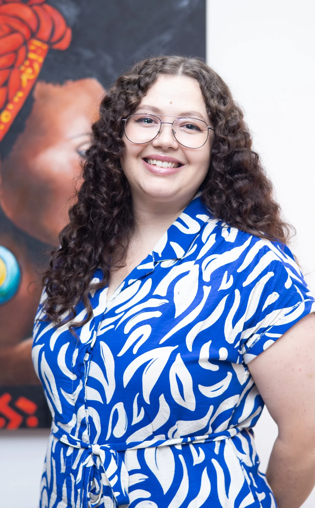 A woman with curly brown hair, wearing glasses and a blue and white patterned dress, smiling at the camera, standing in front of a colorful abstract background.