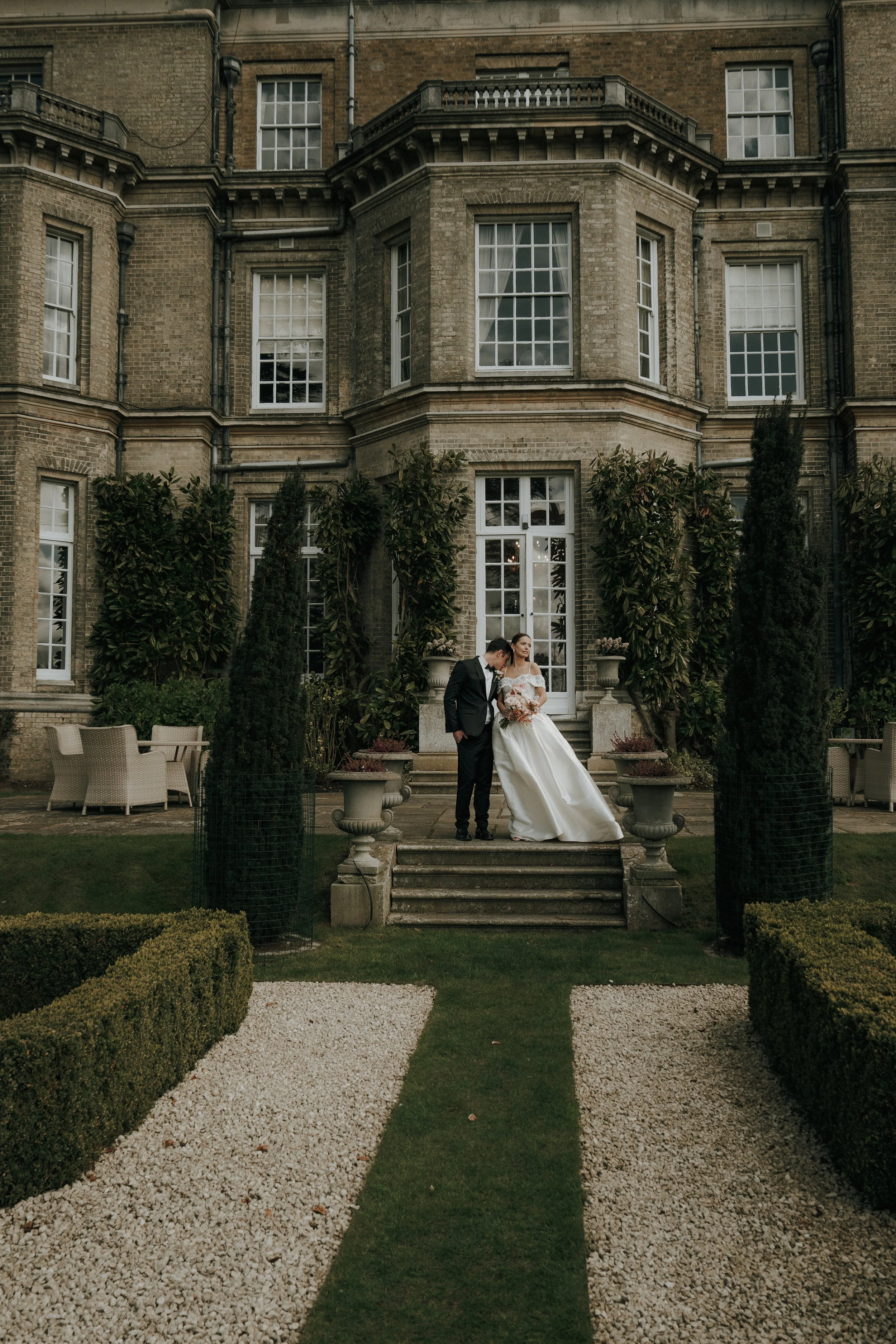 A bride and groom standing on steps in front of a large, historic brick building with tall windows, surrounded by greenery and decorative planters.