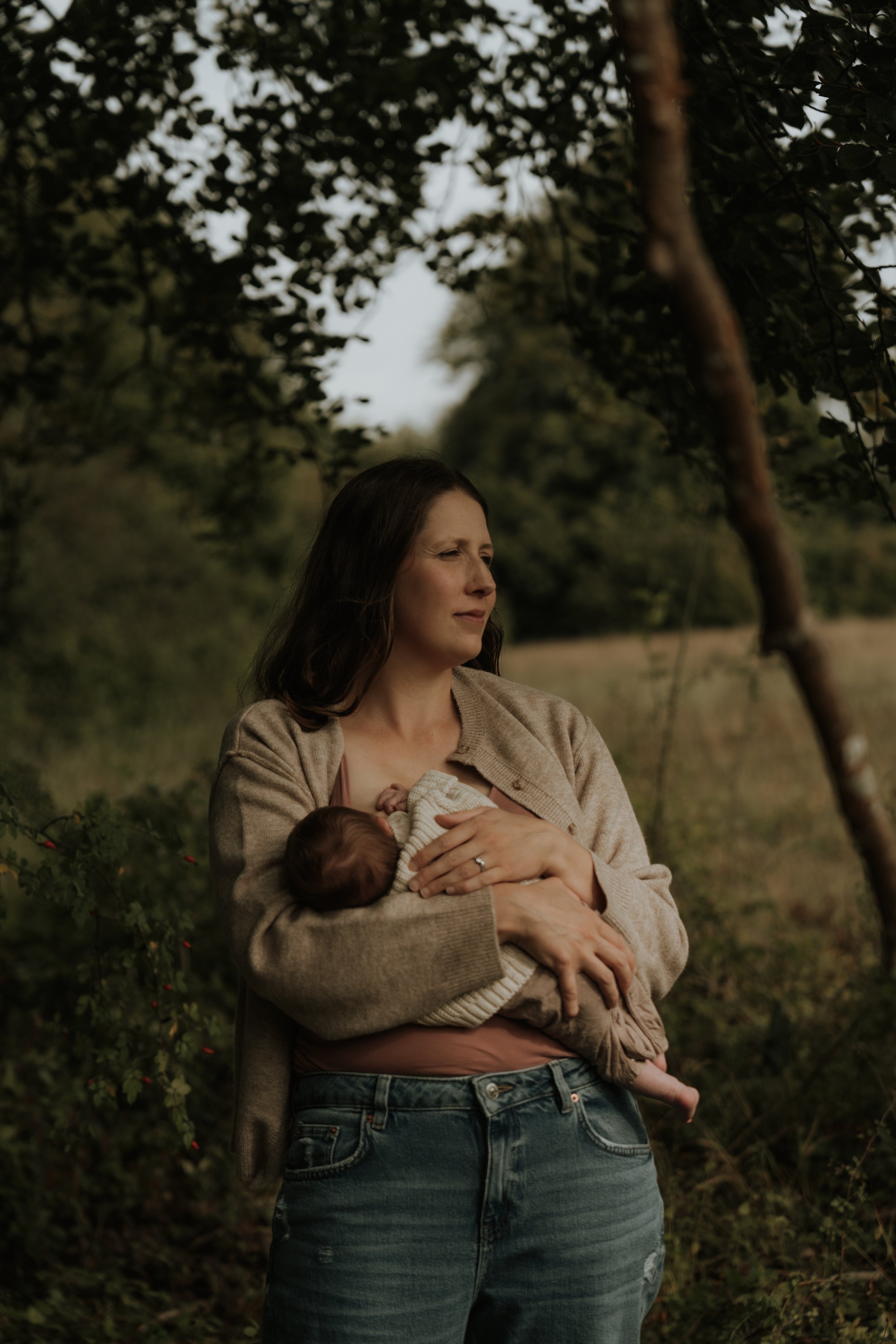 A woman with long brown hair, wearing a beige sweater and jeans, holds a small baby in her arms outdoors amid trees and greenery, with a natural, serene setting.