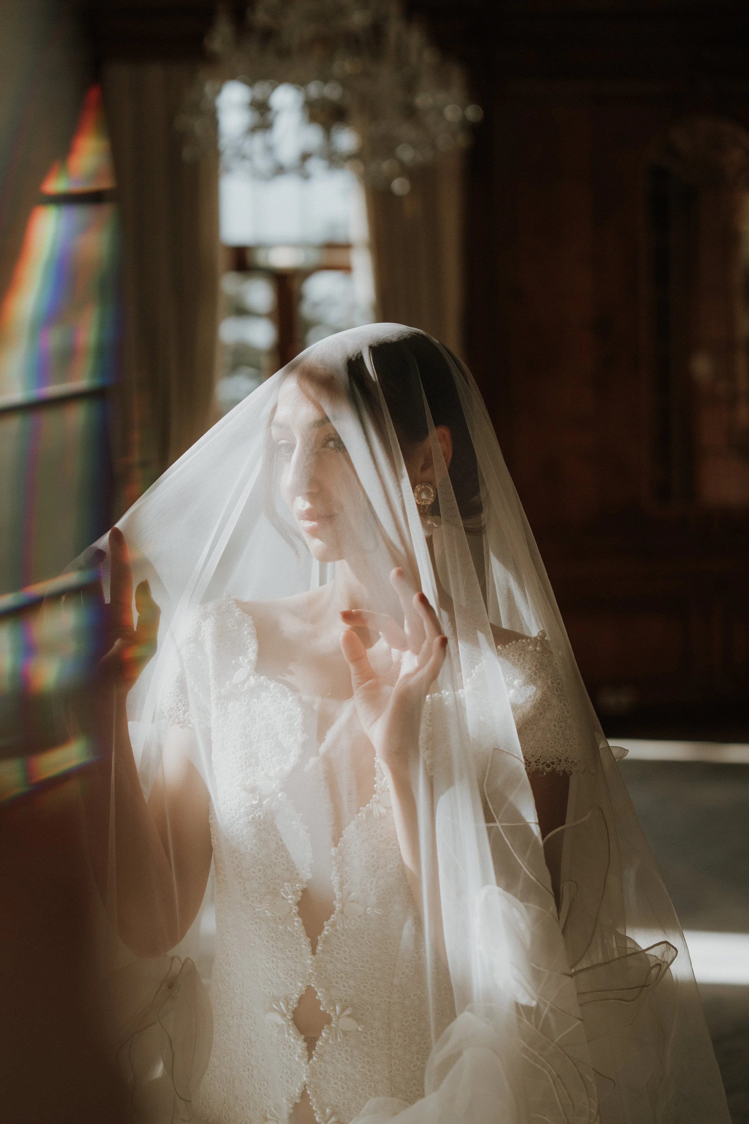 An Editorial bride in a white wedding dress and veil, looking through a window with sunlight streaming in, gently holding her veil with one hand.