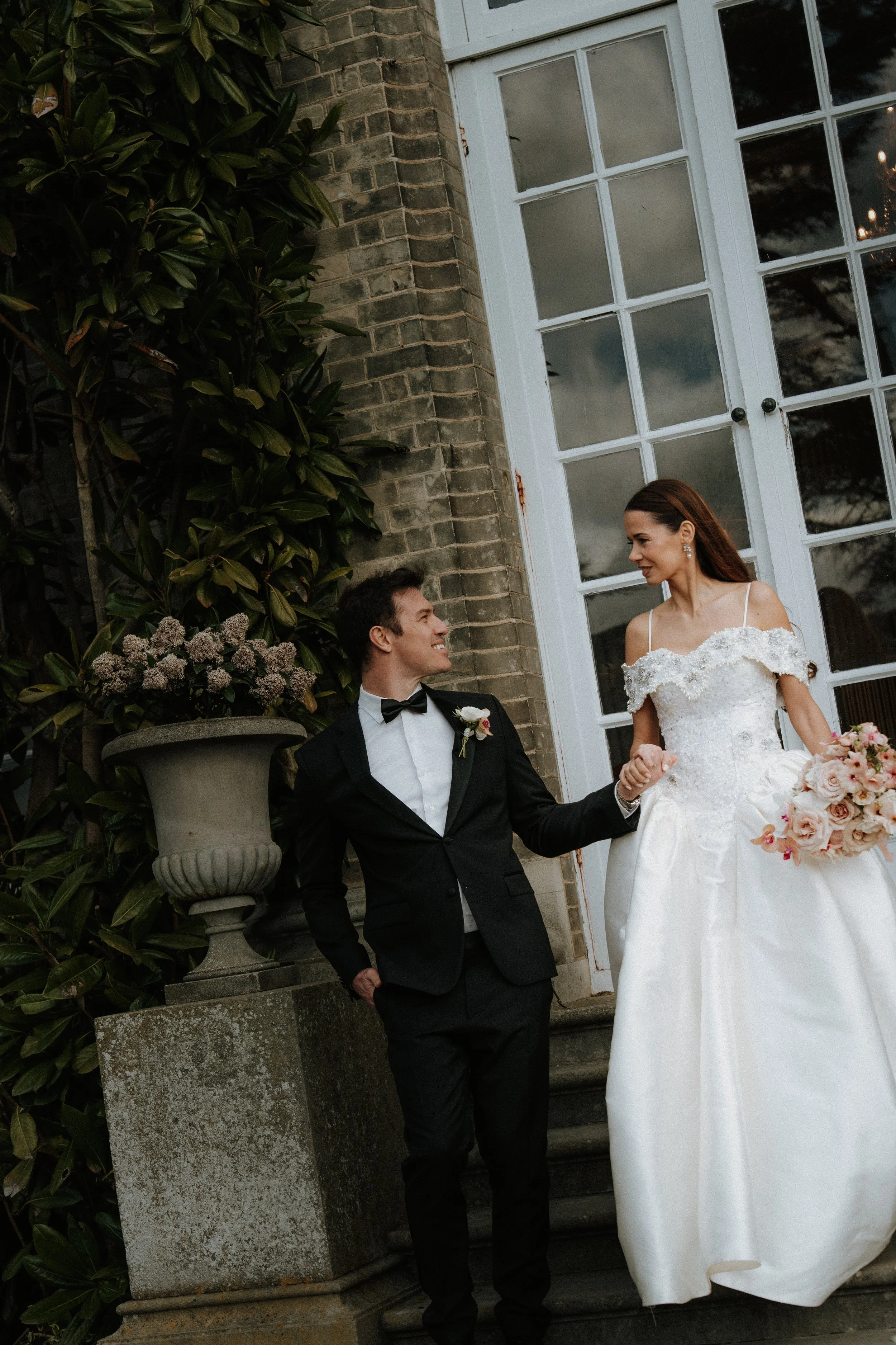 An editorial editorial bride on the steps outside a building, smiling and holding hands. She is wearing a white wedding dress with off-the-shoulder sleeves and holding a bouquet of pink flowers. The groom is wearing a black tuxedo with a bow tie.