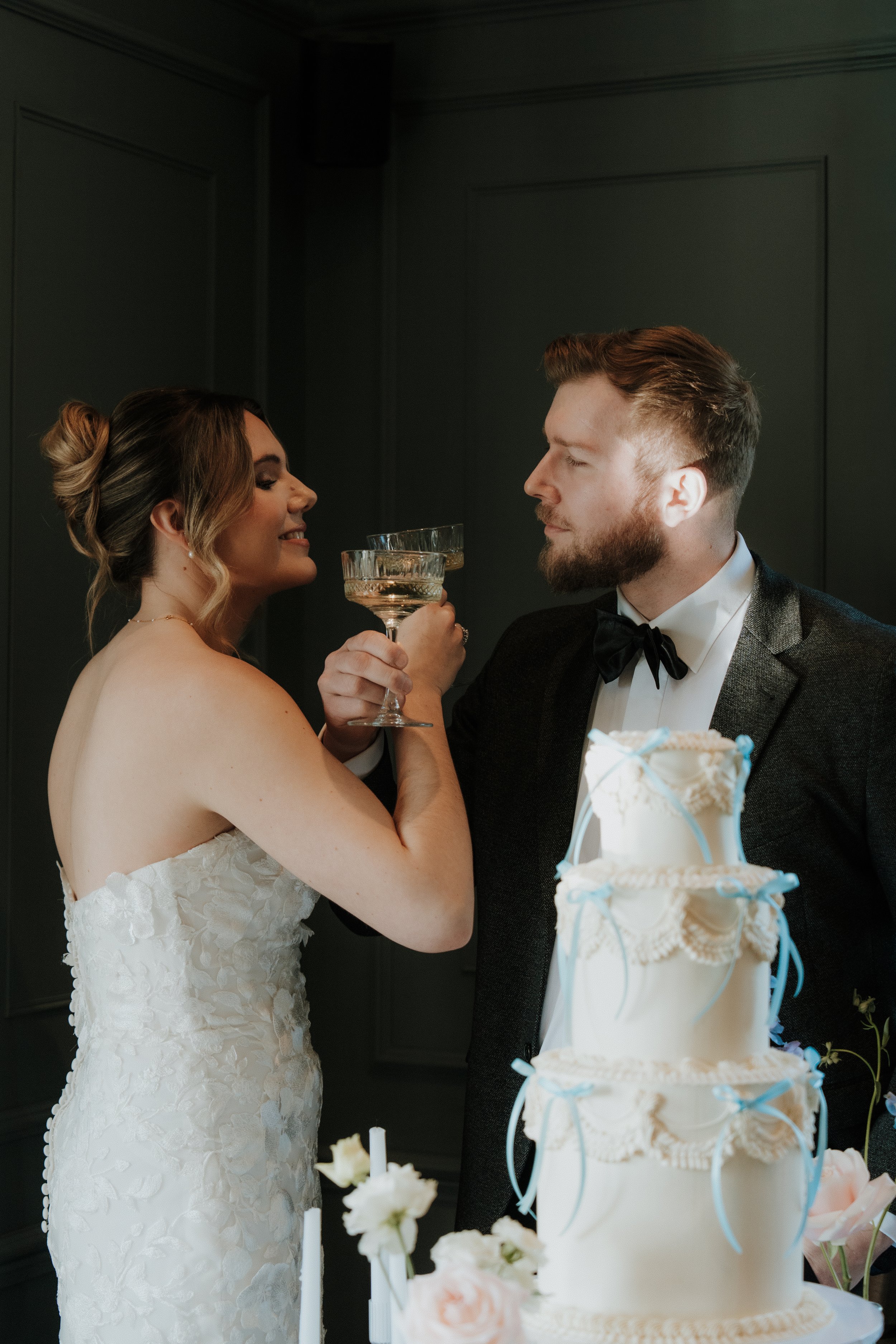 Bride and groom at their wedding celebration, holding champagne glasses and smiling at each other, with a wedding cake in front of them.