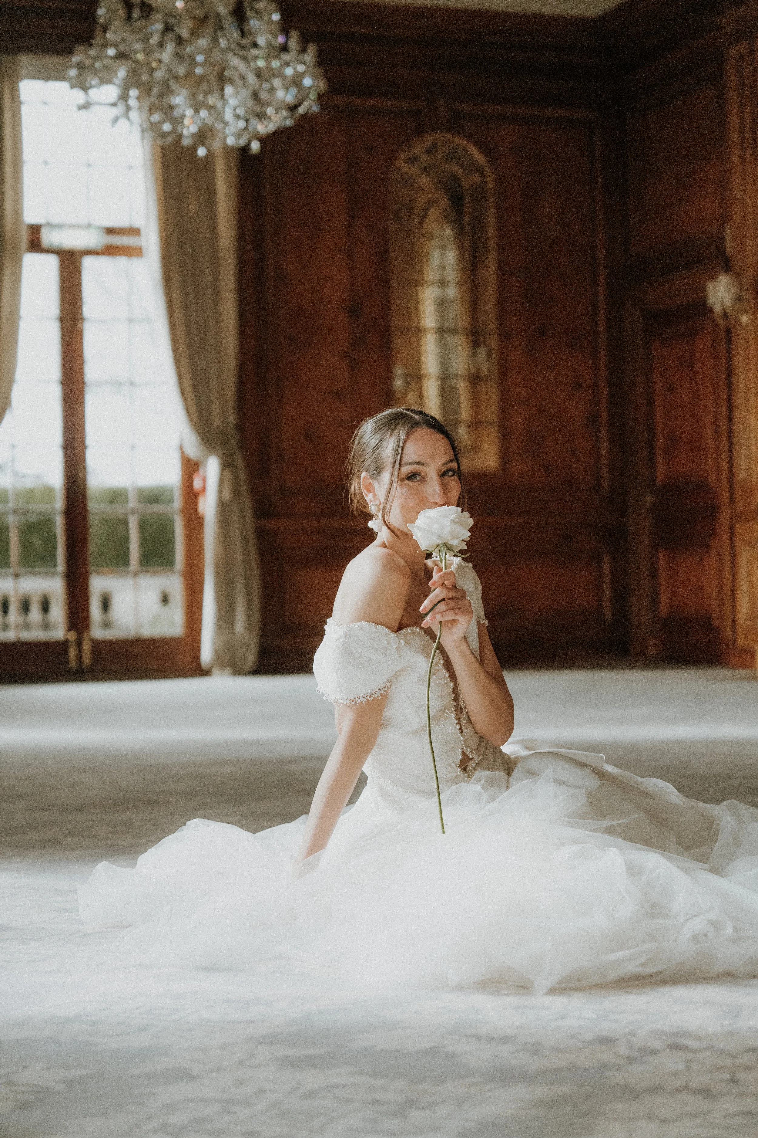 An Editorial bride sitting on the floor in a wedding gown, holding a white rose near her face inside a grand, wood-paneled room with large windows and a chandelier.