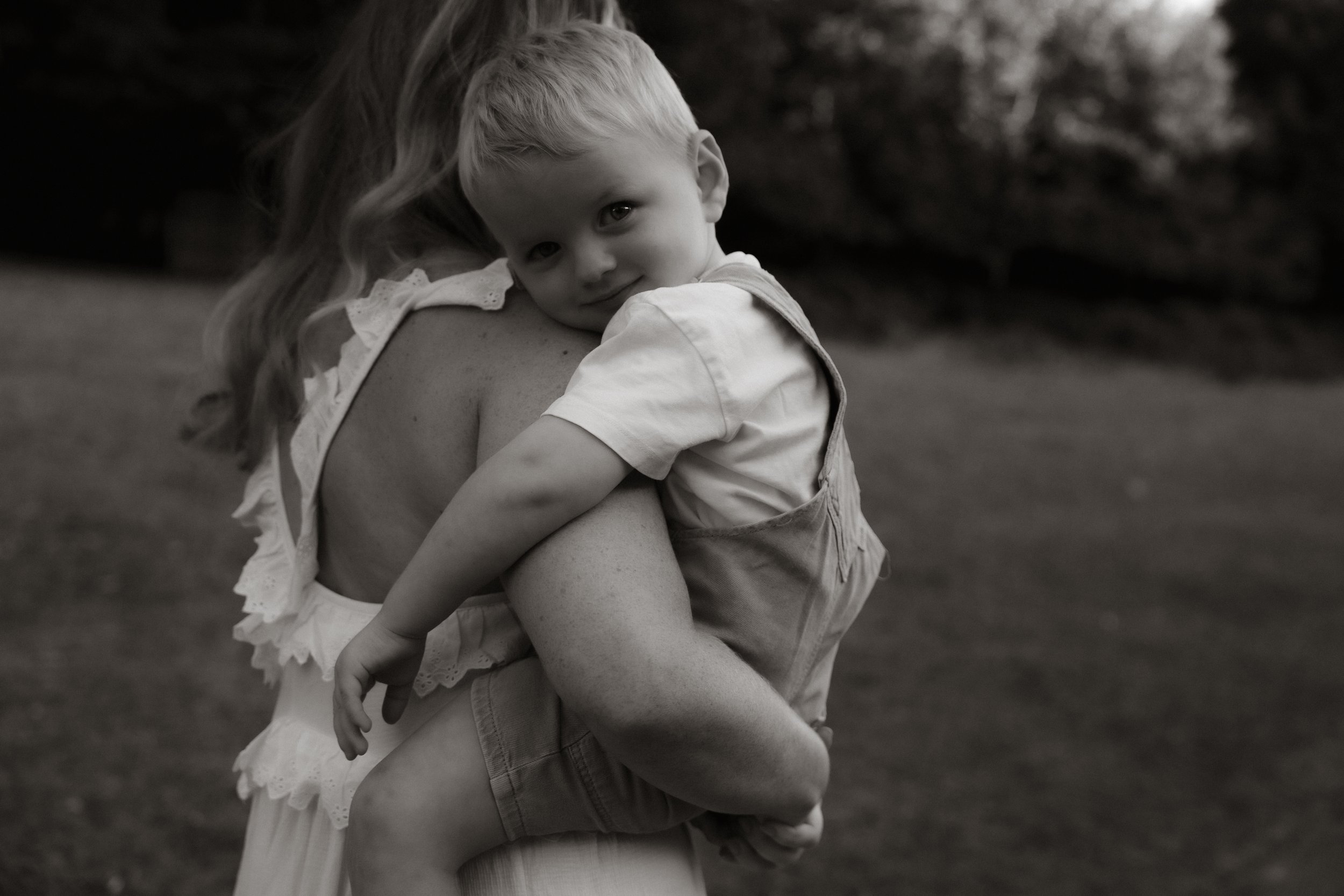 A young child with short blonde hair and a girl with long hair, hugging outdoors. The child looks at the camera, while the girl is not fully visible.