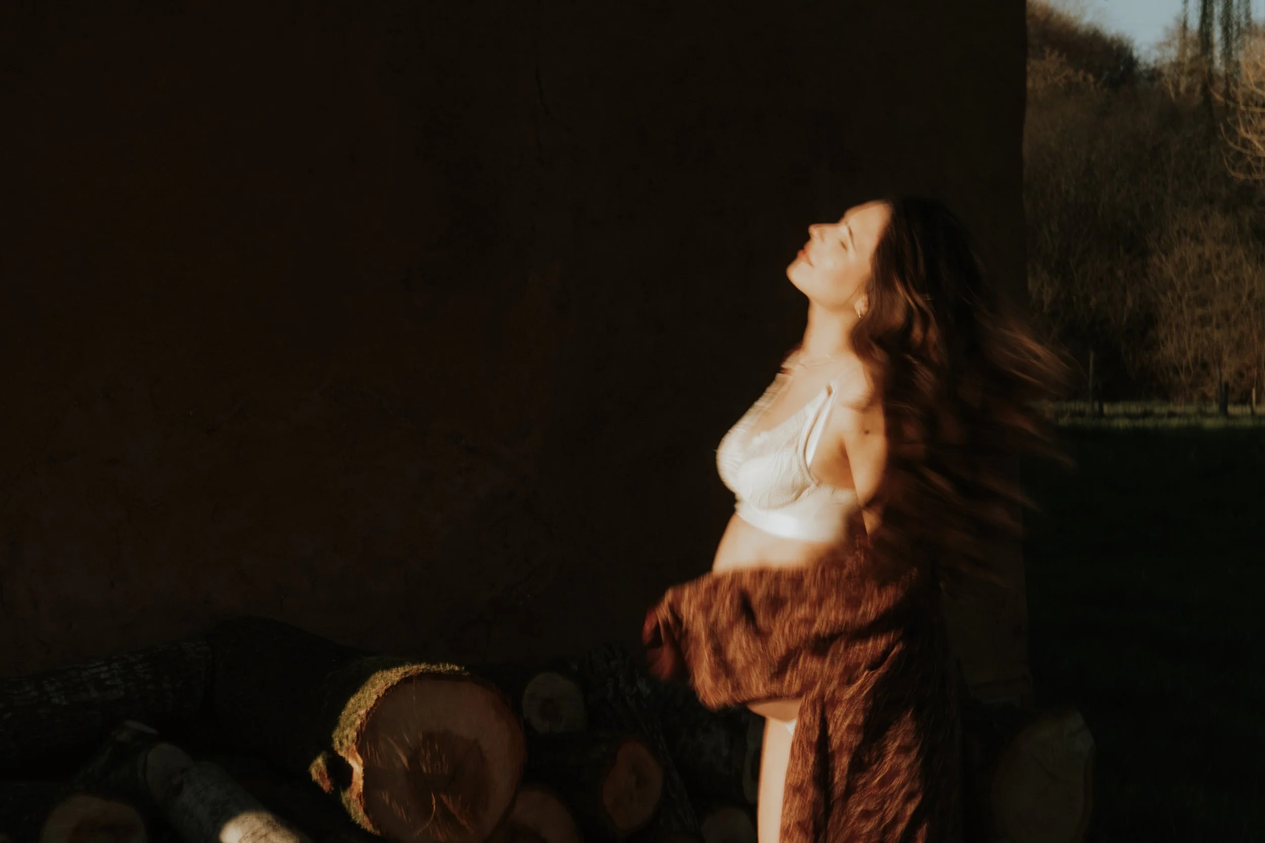 Editorial maternity shoot - A woman standing outdoors at sunset with her eyes closed, facing the sunlight. She is wearing a white top and has long, flowing hair. There is a stack of logs beside her.