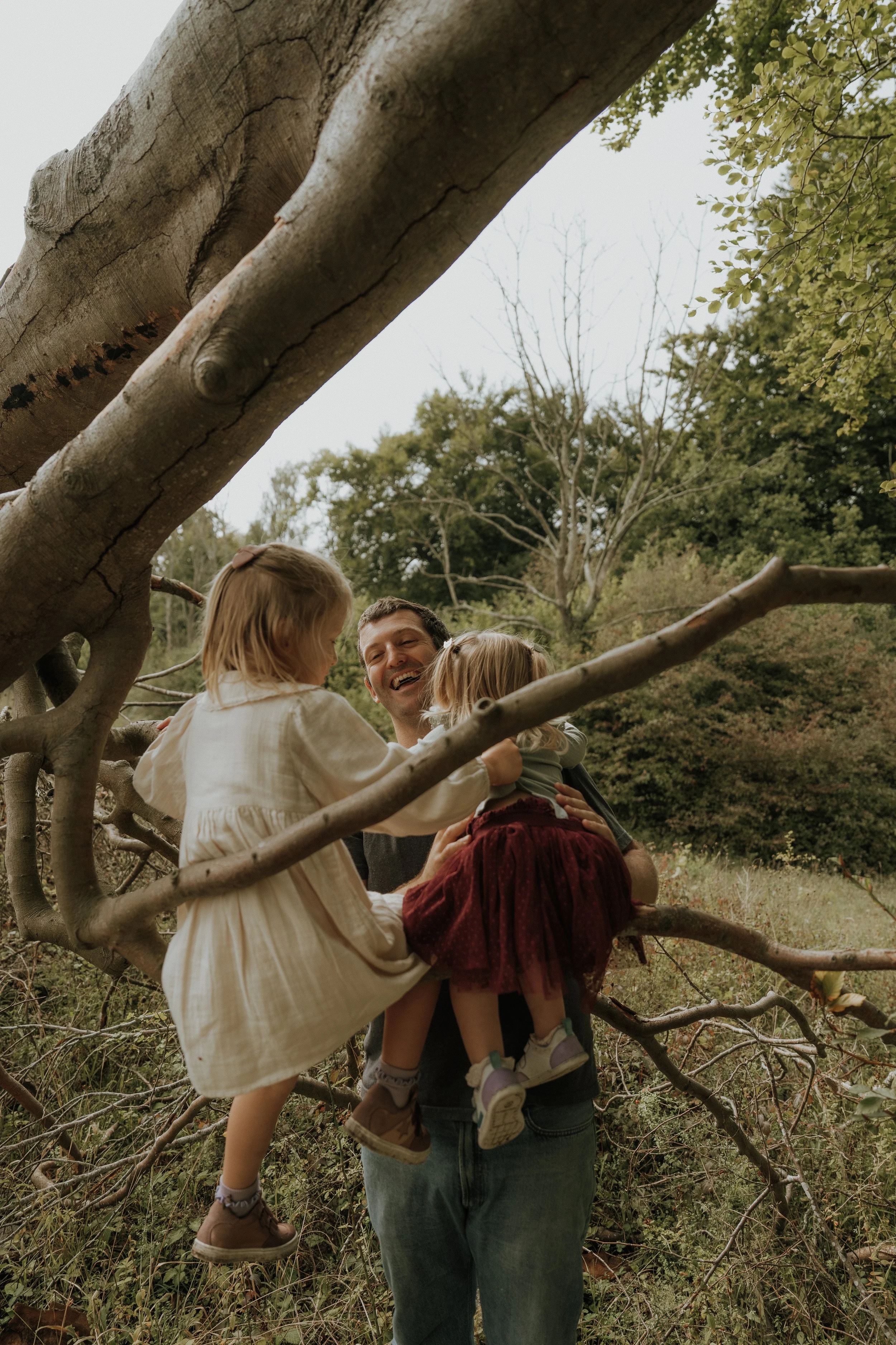 A father holding two young children, one on each arm, while they sit on tree branches in a forested area with trees and foliage in the background.