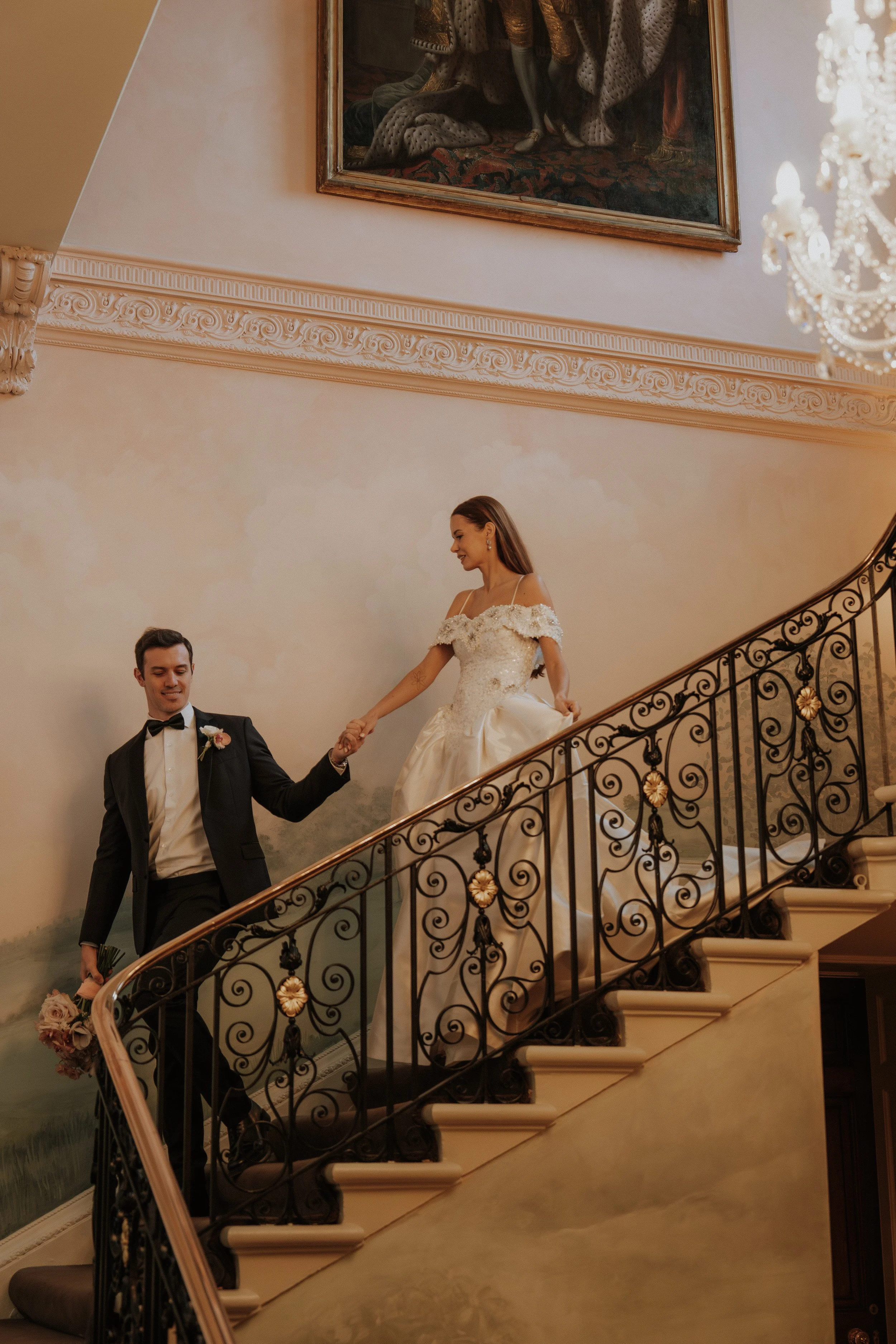 A newlywed couple is ascending an ornate staircase inside an elegant building. The groom is dressed in a black tuxedo and holding a bouquet of flowers, while the bride is wearing a white wedding gown and smiling as she holds the groom's hand.