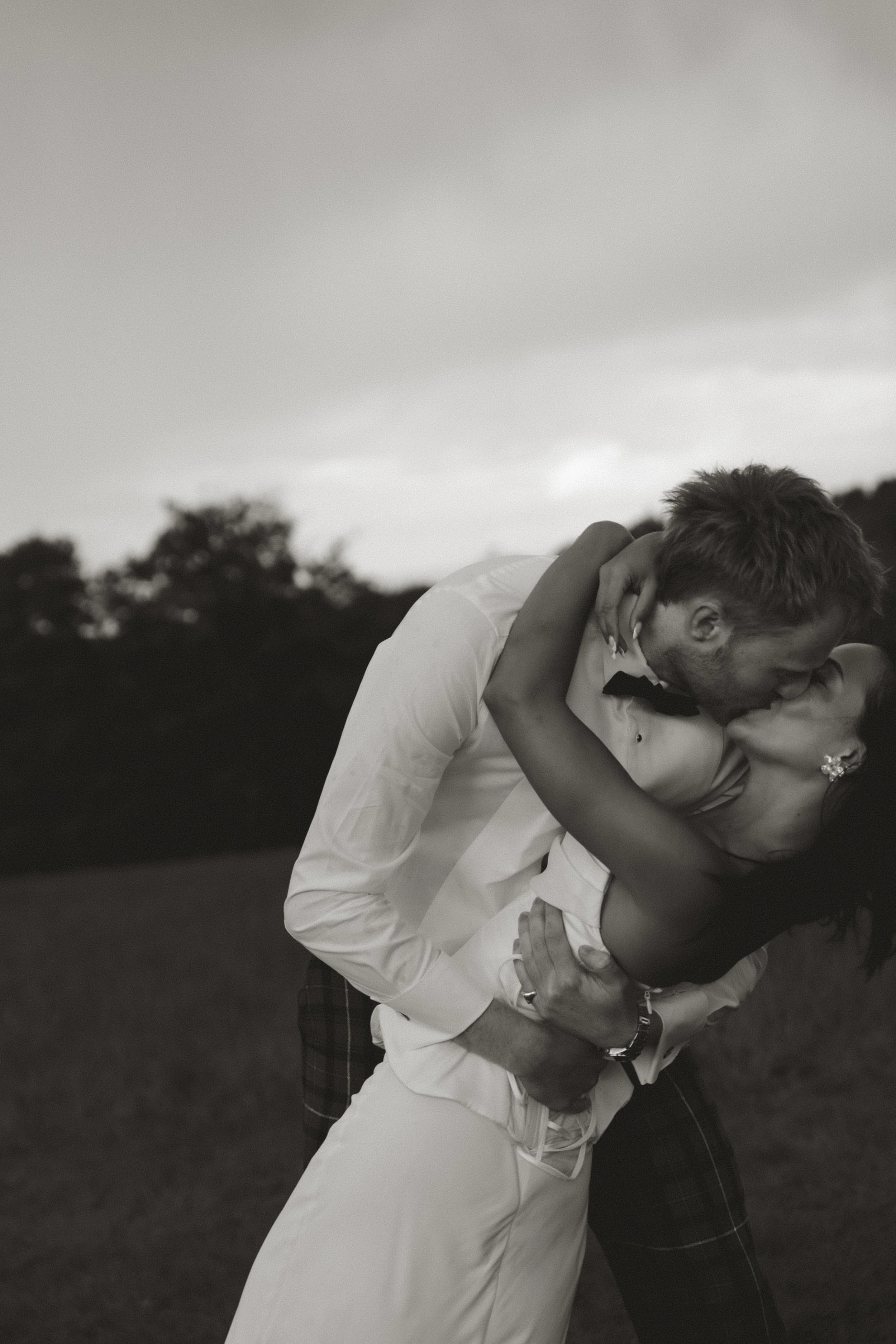 A couple sharing a dance outdoors, with the woman in a white dress and the man in a white shirt and bow tie, their faces close together.