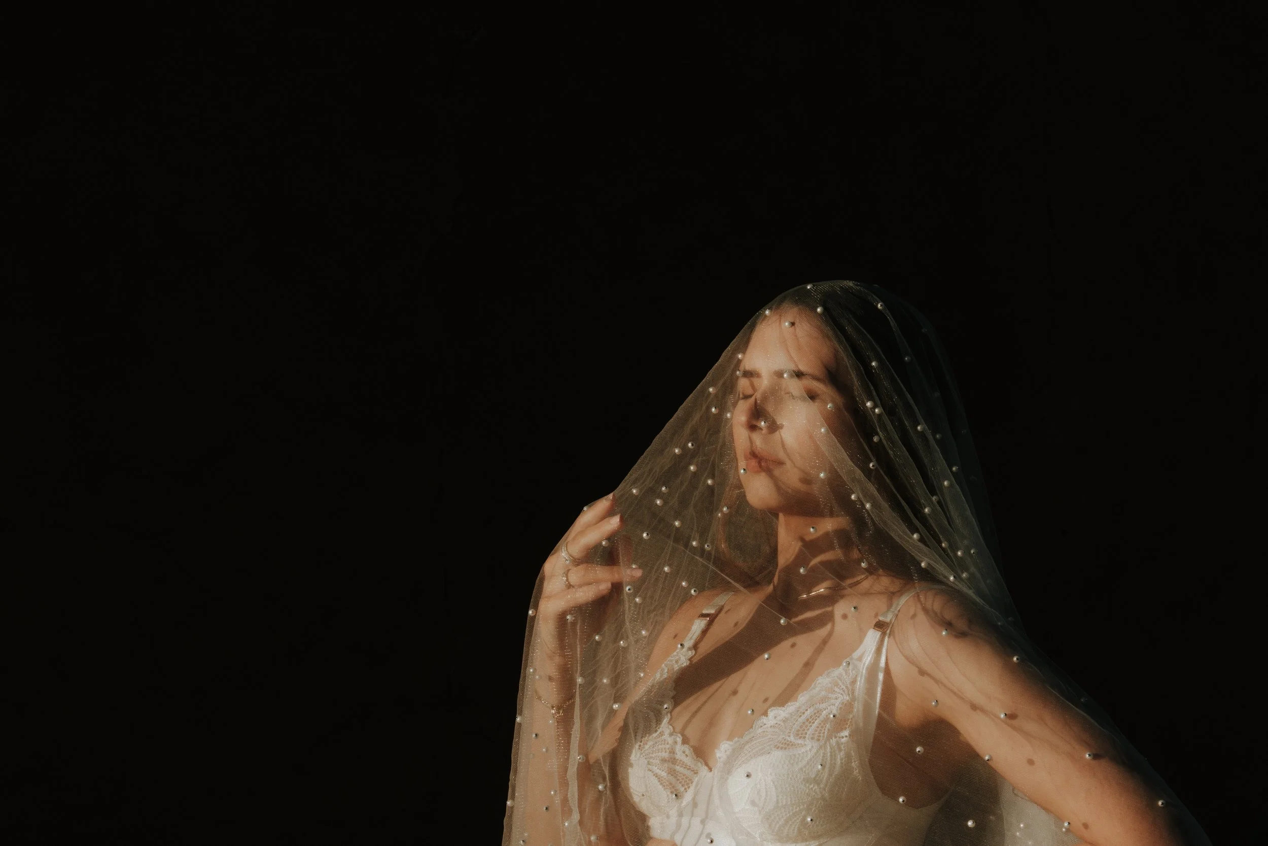 Editorial maternity shoot - A woman with closed eyes wearing a white lace dress and a veil with pearl embellishments on a dark background.