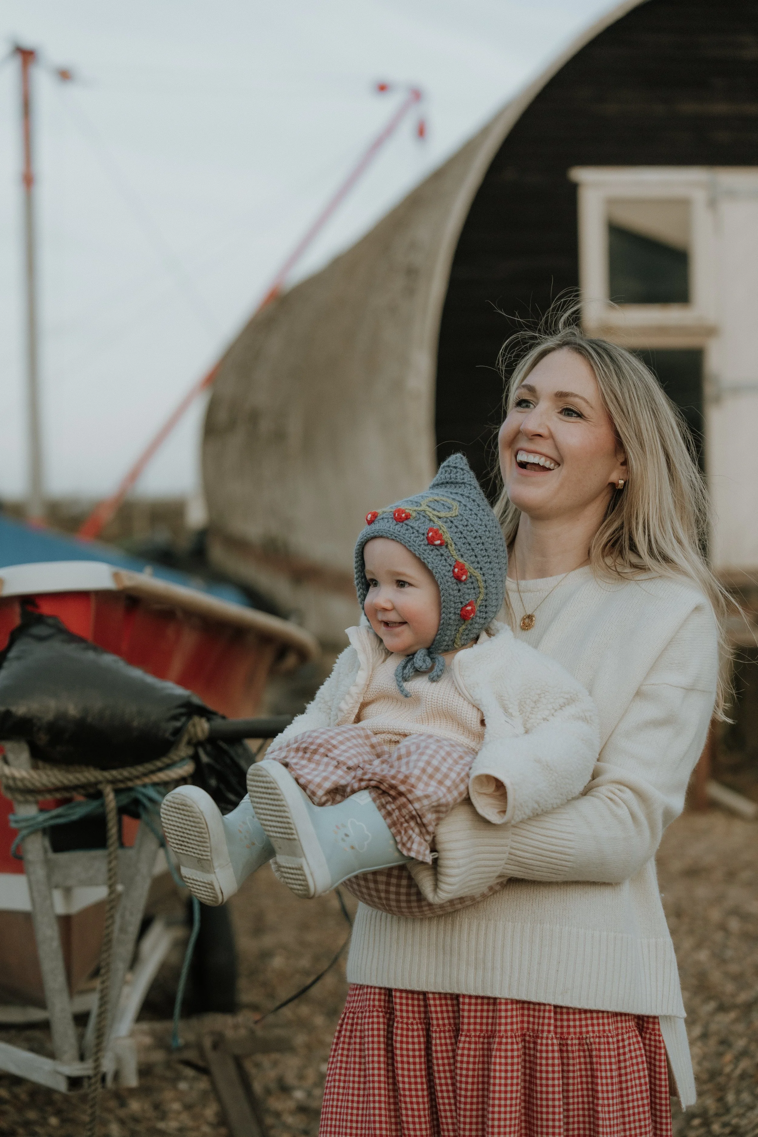 A mother holding a smiling child wearing a knit hat with cherries, outside near boats and a curved wooden structure, during daytime.