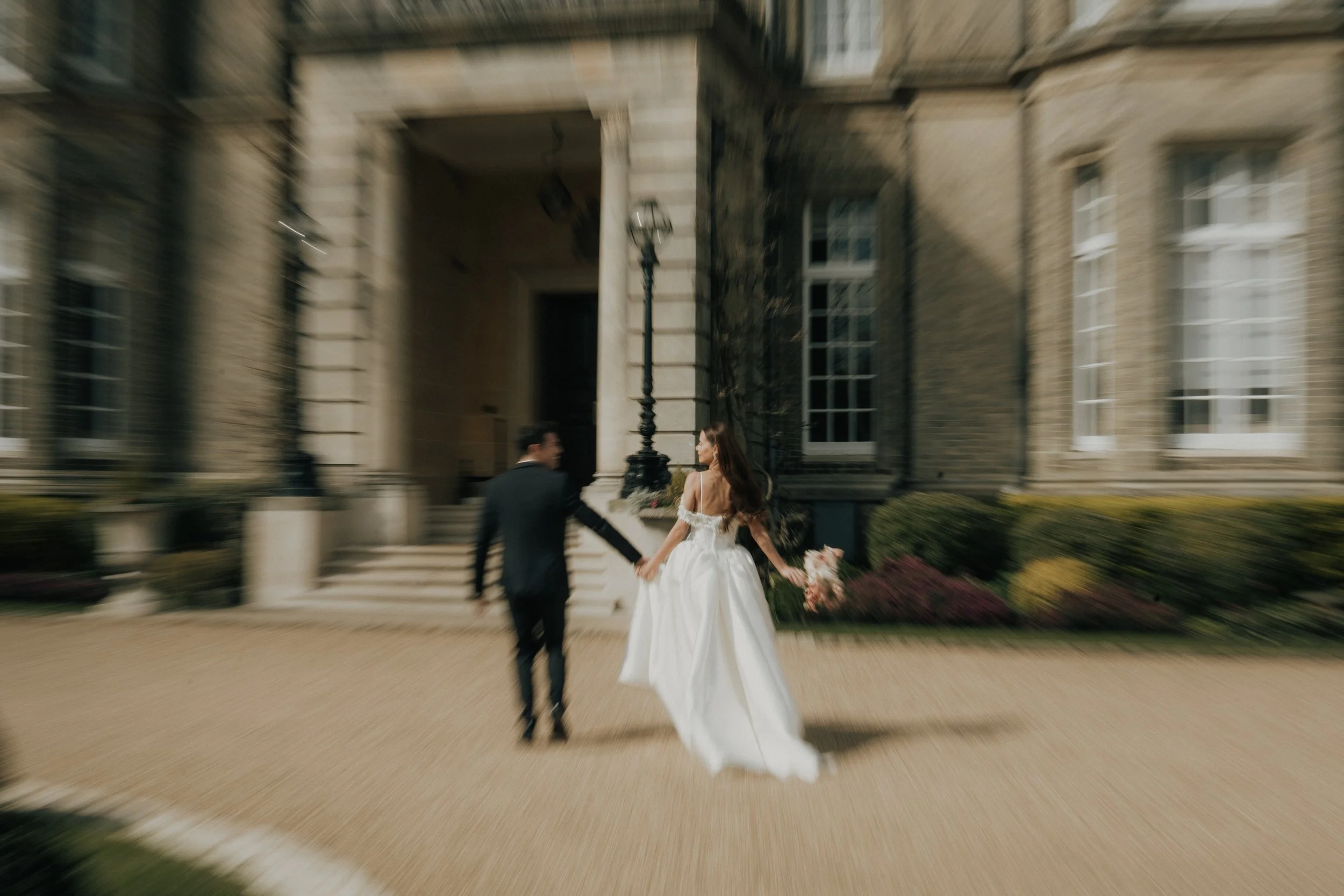 An editorial editorial bride and groom holding hands and walking away from a grand stone building, Hedsor House, with stairs and windows, on a sunny day.