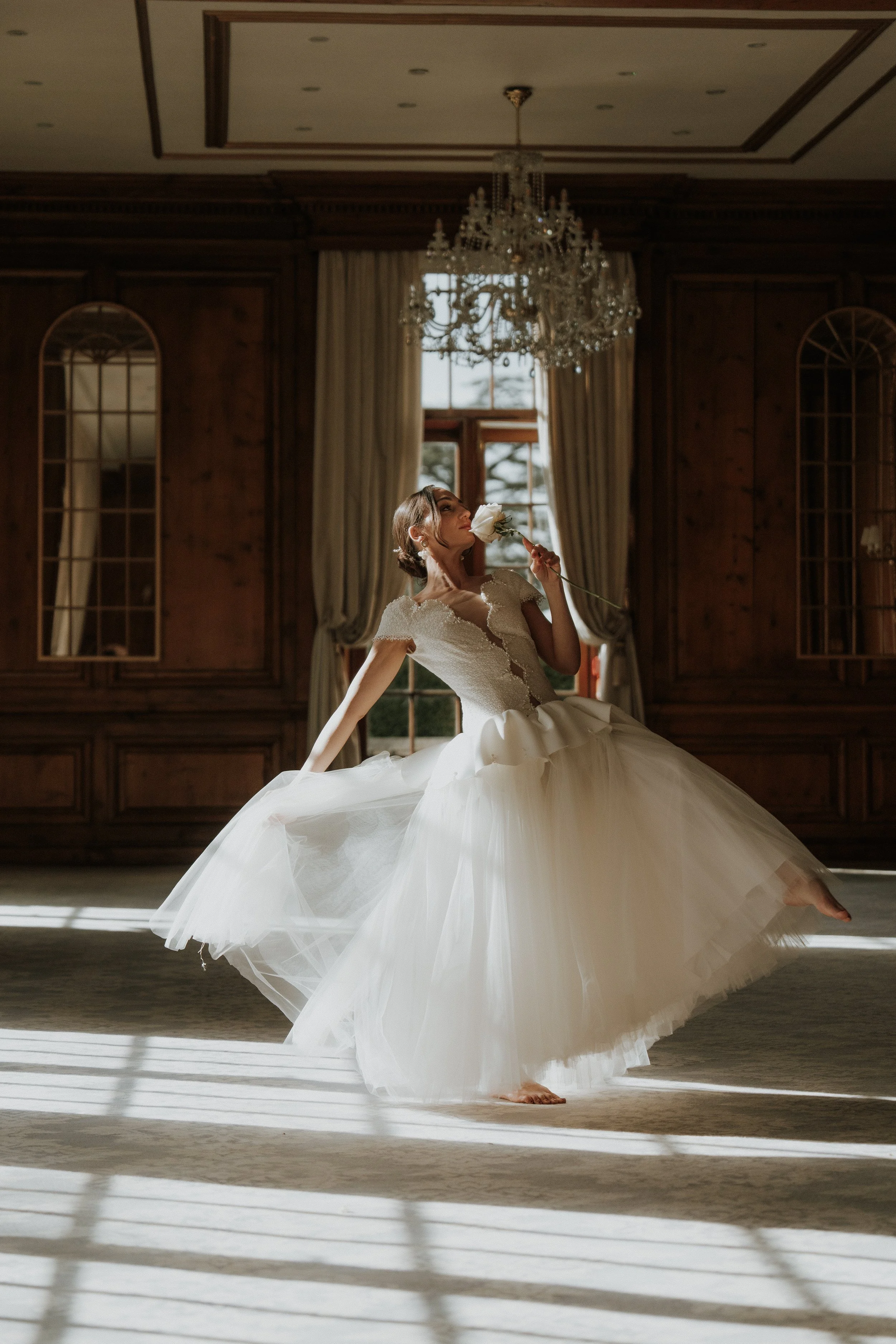 An Editorial Bride in a wedding dress dancing barefoot in a large, elegant room with dark wood paneling, a chandelier, and tall windows with curtains, holding a white rose near her face.