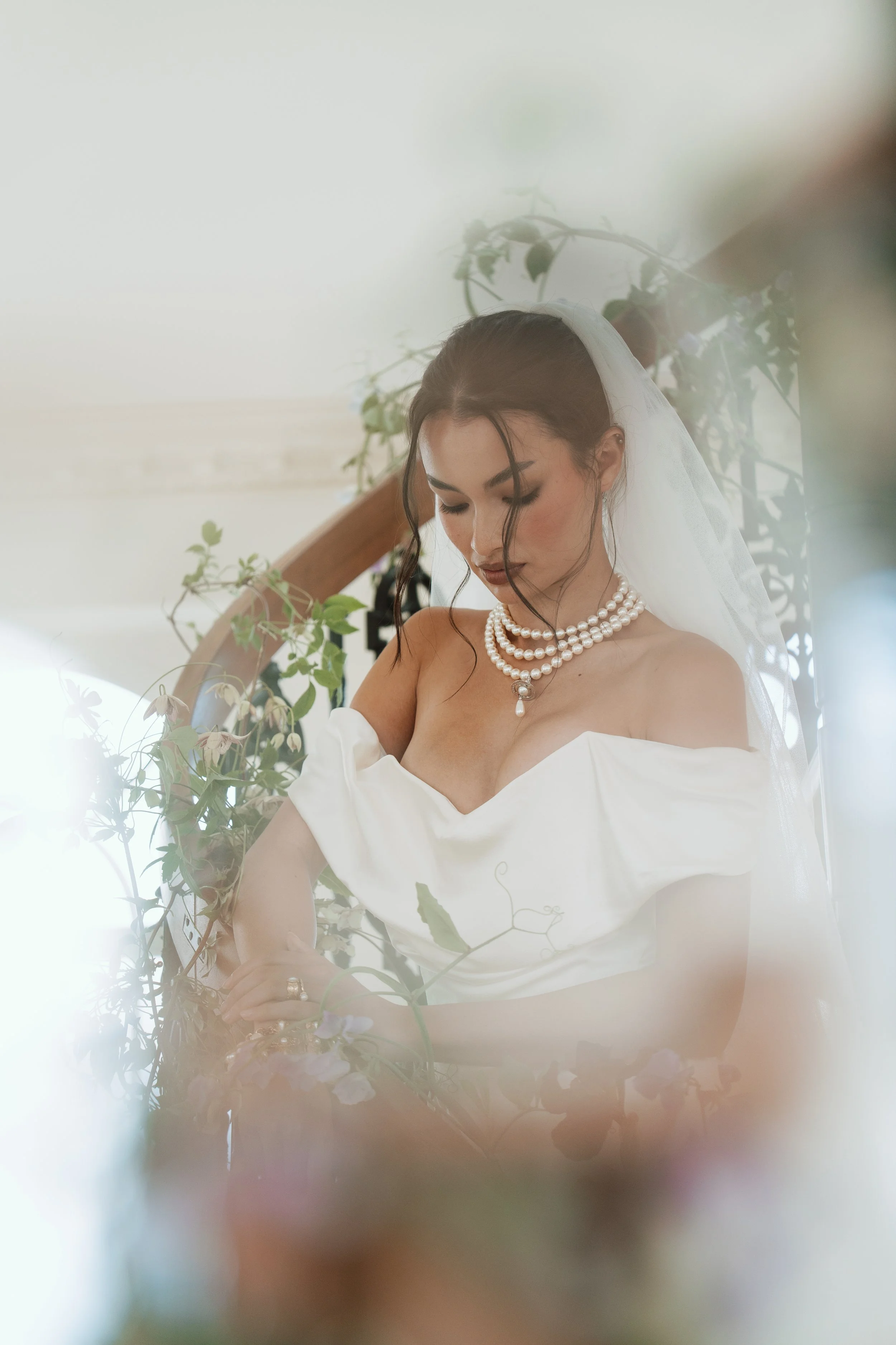 A bride with dark hair styled in an elegant updo, wearing pearl necklaces, a white off-shoulder wedding dress, and a veil, standing amidst greenery and flowers.