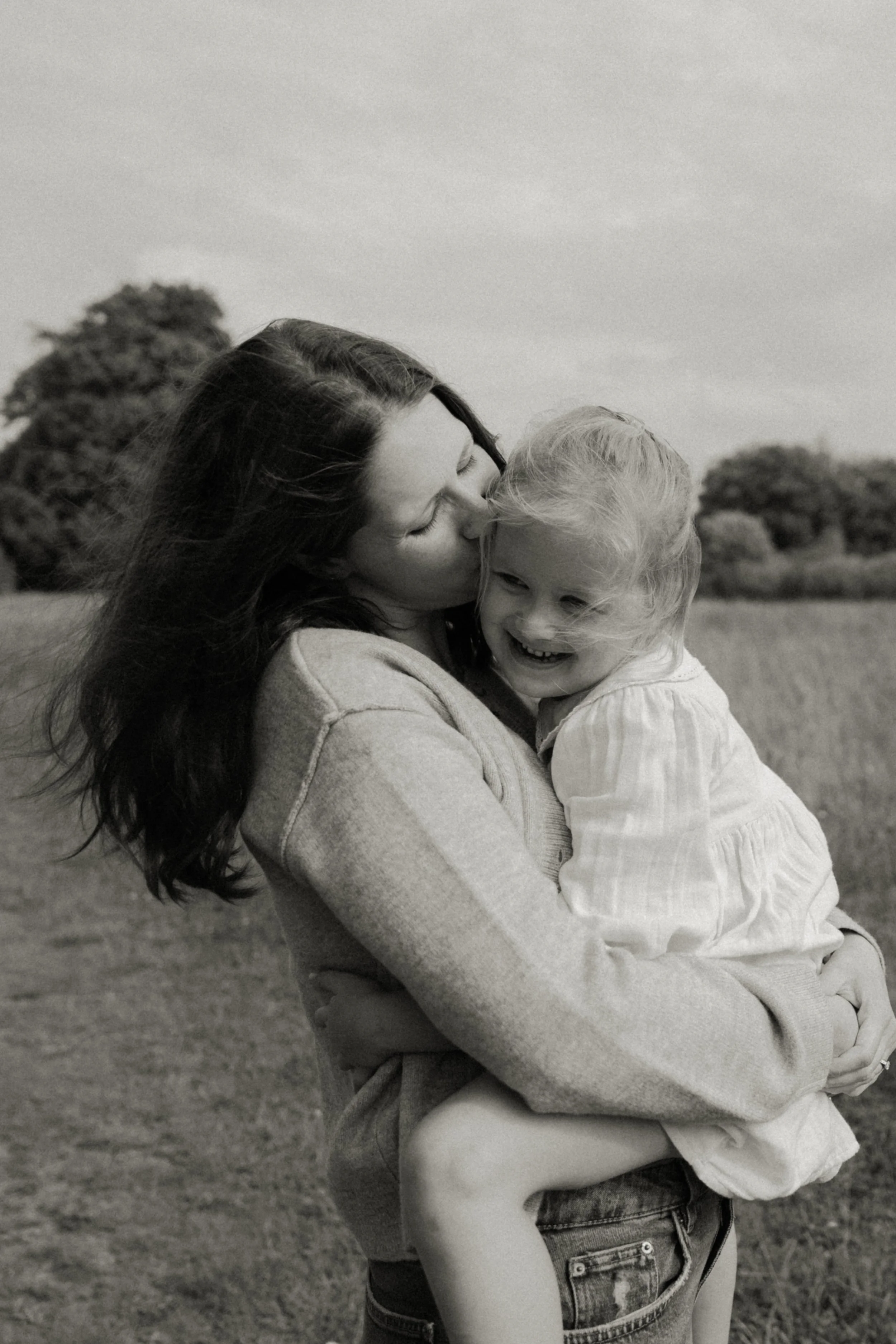 Black and white photo of a woman holding a young girl, kissing her on the cheek in a park with trees in the background.