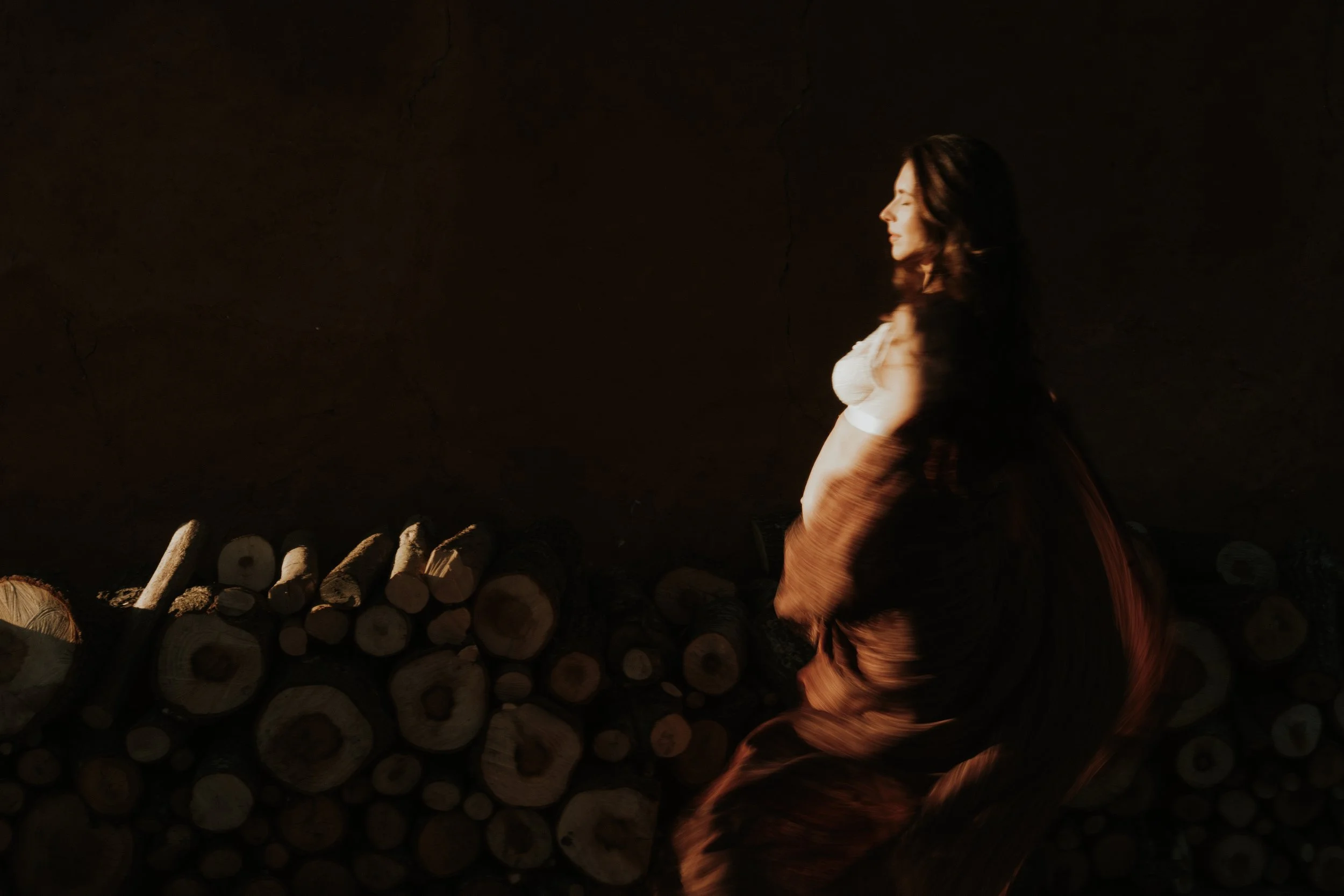 Editorial maternity shoot - A woman with long dark hair, wearing a white crop top and a flowing skirt, stands sideways next to a pile of cut logs against a dark background.