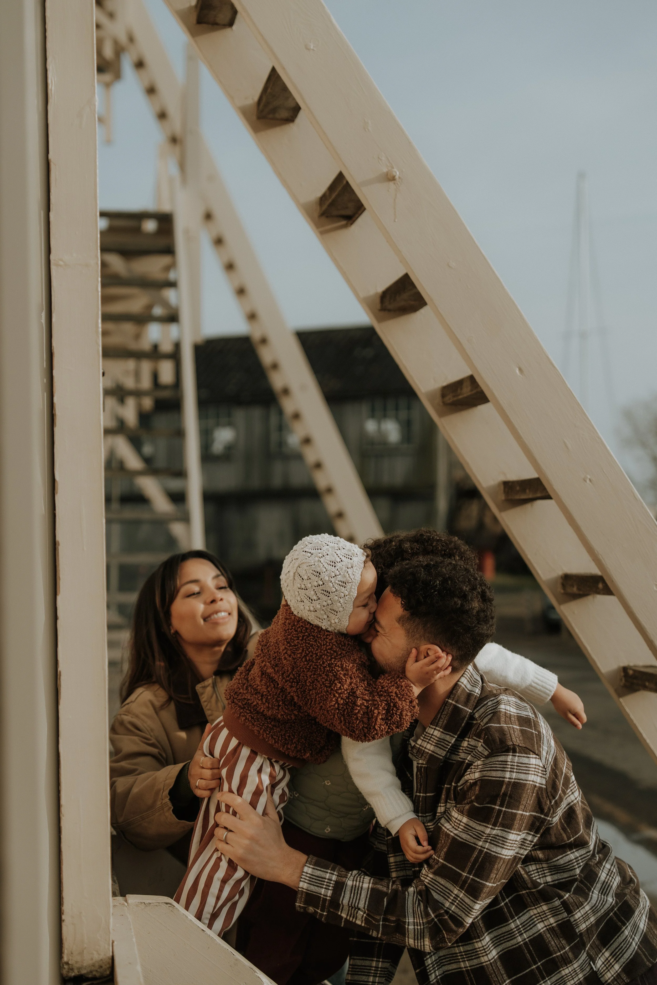 A family outdoors on a staircase, with a woman, a man, and two young children, one of whom is kissing the man on the nose.