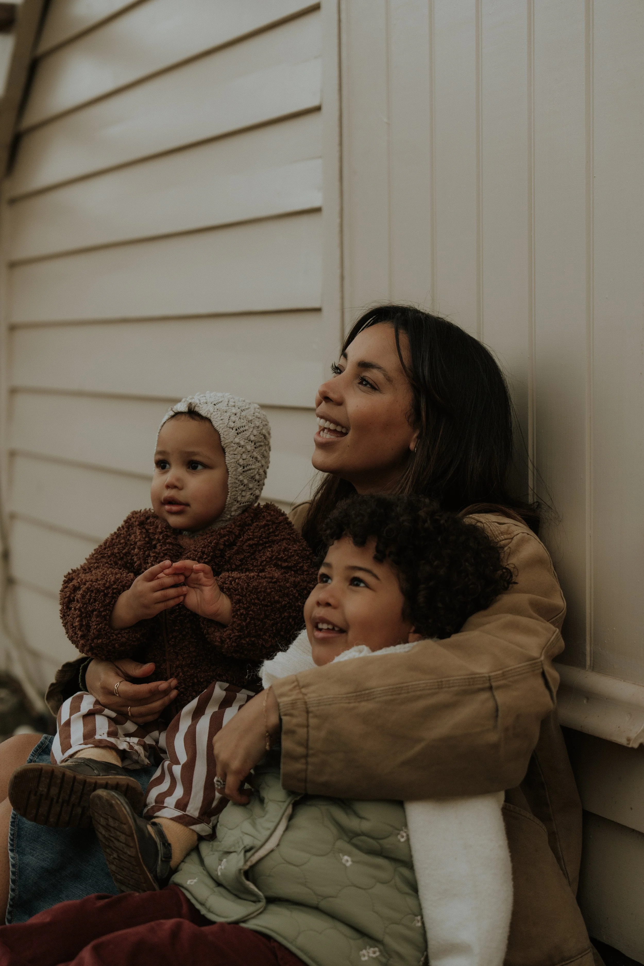 A mother sitting against a white wall, smiling while holding two young children, one with curly hair and the other in a knit hat, all dressed warmly.