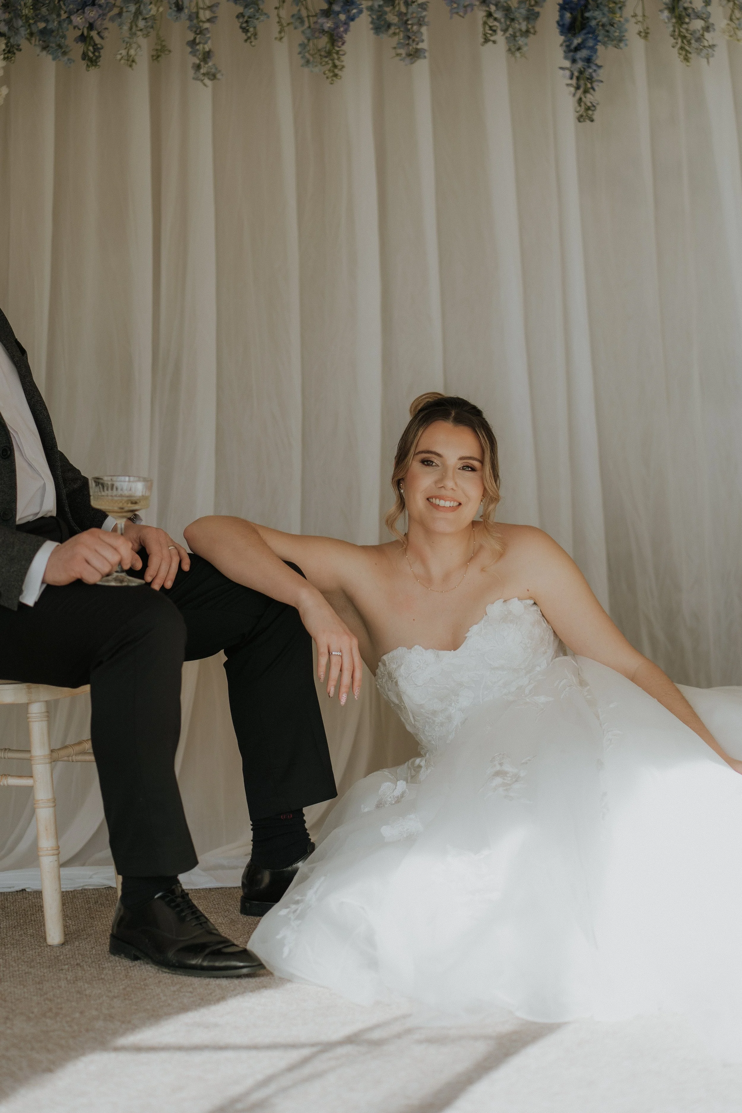 Bride in a white wedding gown sitting on the floor, smiling, with a man sitting beside her holding a drink, in front of a cream-colored curtain backdrop decorated with hanging purple and green flowers.