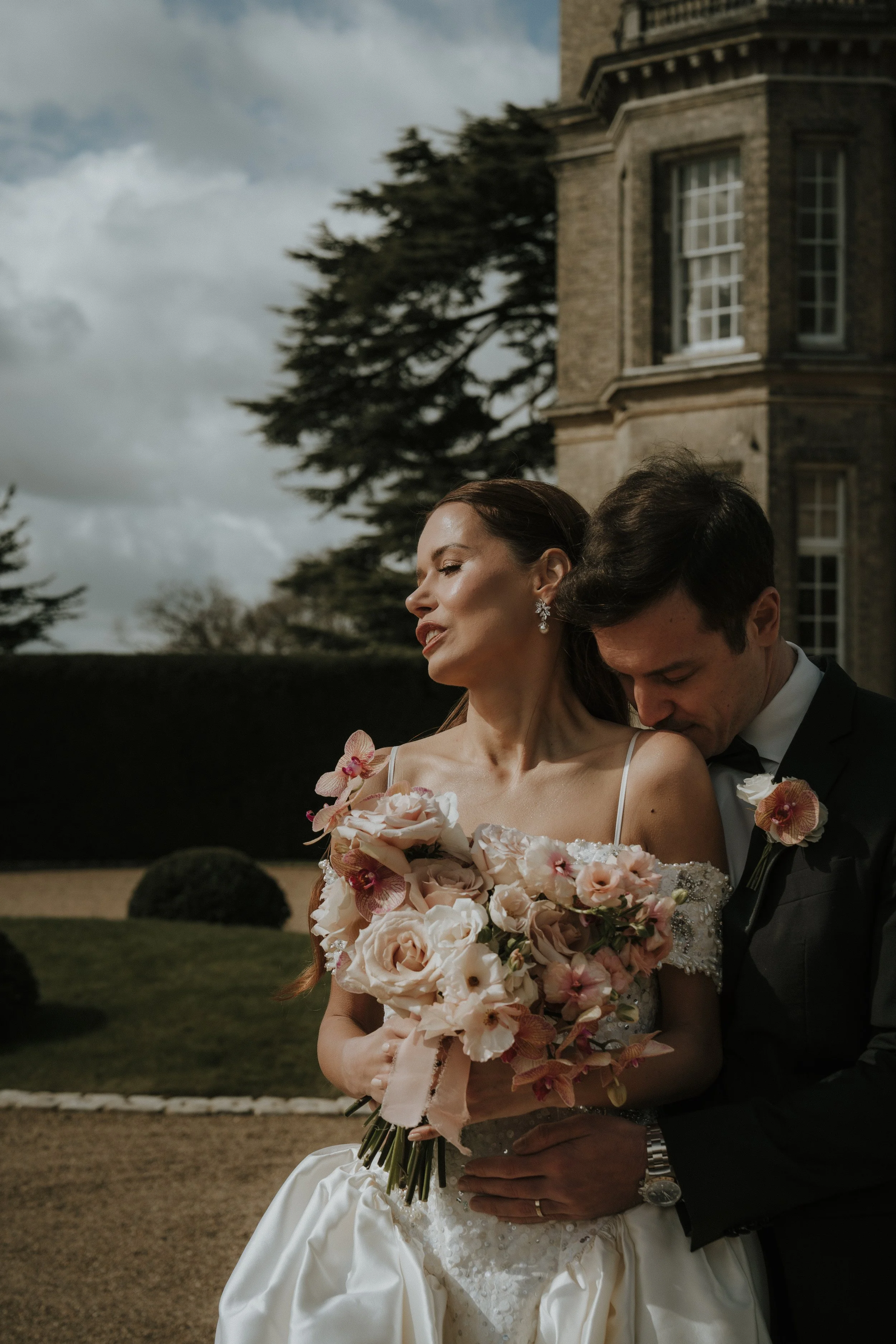 An editorial bride and groom on their wedding day outdoors, with the bride holding a bouquet of pink and peach flowers and the groom hugging her from behind, in front of a historic stone building and trees.