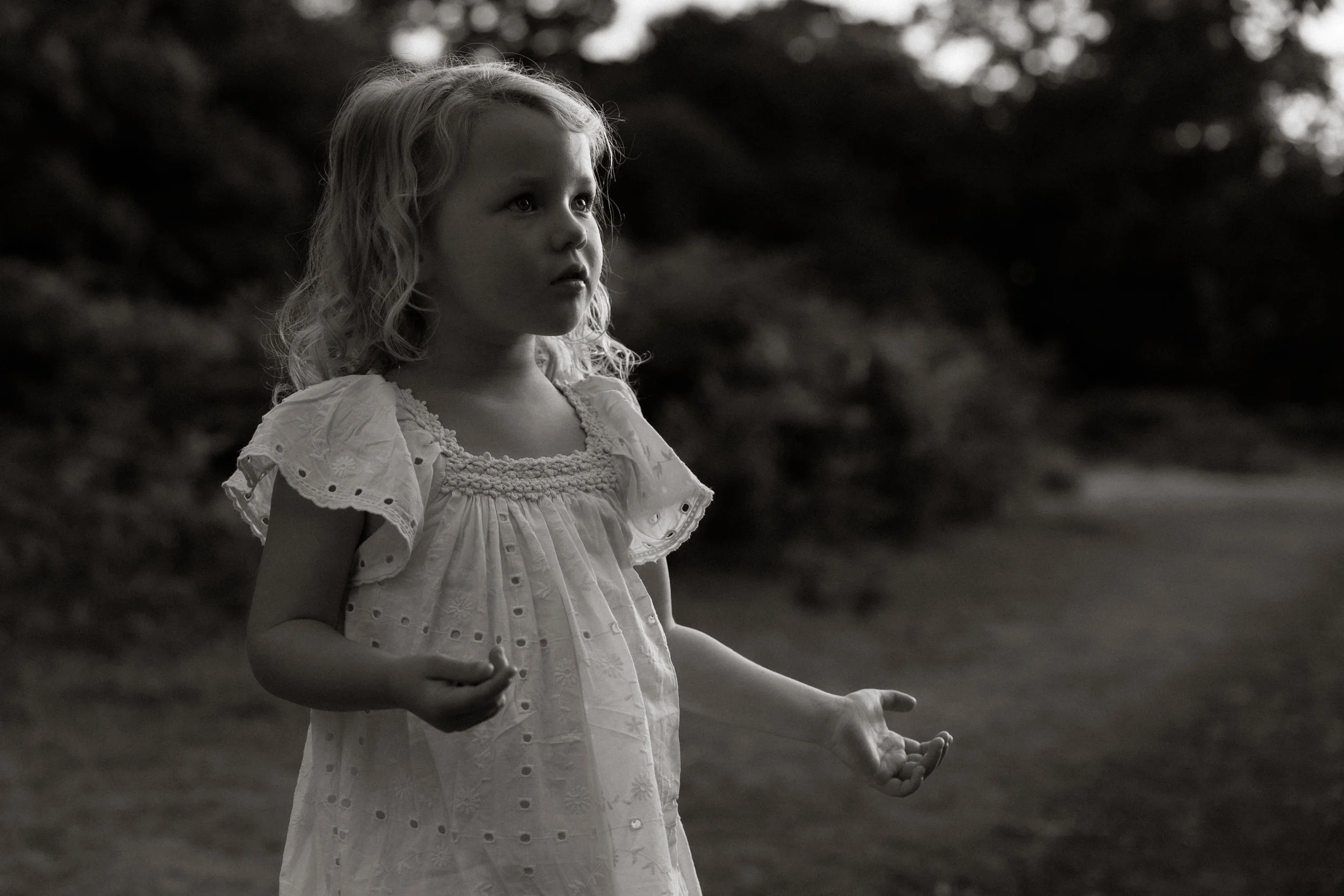 A young girl with curly hair wearing a white dress stands outdoors in a black and white photo, looking pensively to the side with her hands held slightly open.