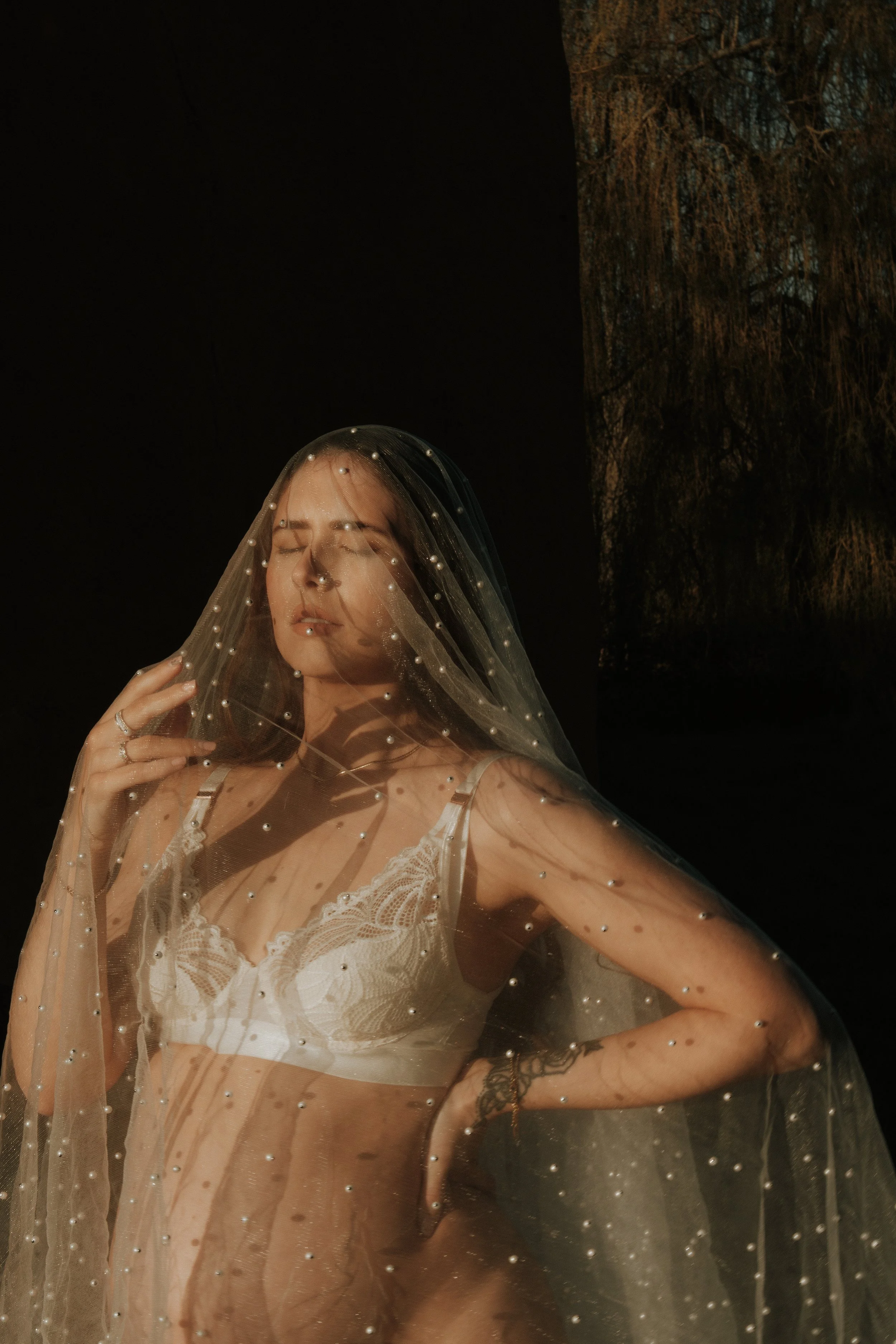 Editorial maternity shoot - A woman with closed eyes wearing lace lingerie and a pearl-adorned veil, standing outdoors with shadows cast on her, at dusk or night.