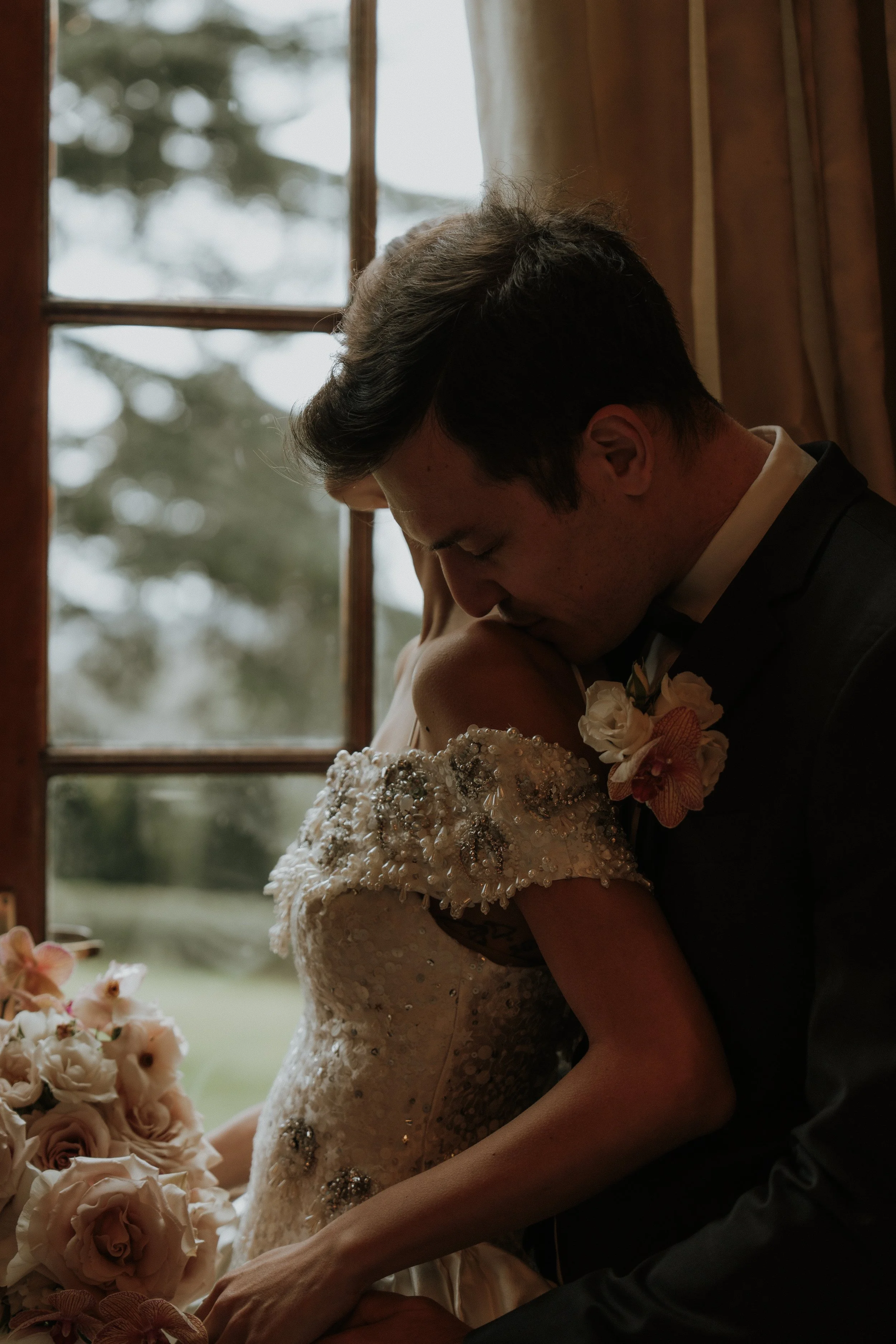 An editorial editorial bride and groom in wedding attire sharing an intimate moment indoors near a window, with a blurred outdoor scene in the background.