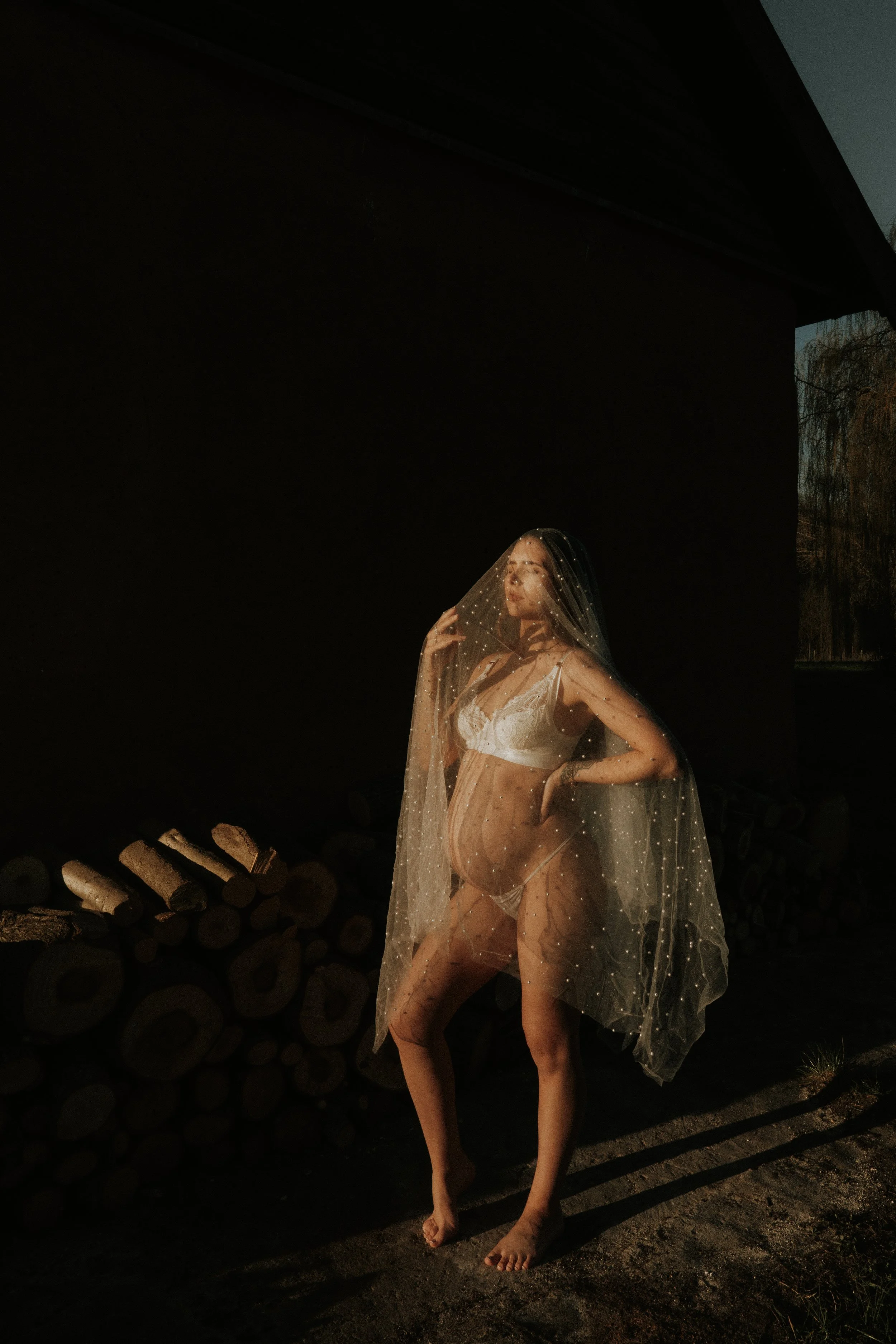 Editorial maternity shoot - A woman in lingerie standing outdoors, wearing a sheer veil with small pearls, with firewood stacked behind her, and sunlight casting shadows.
