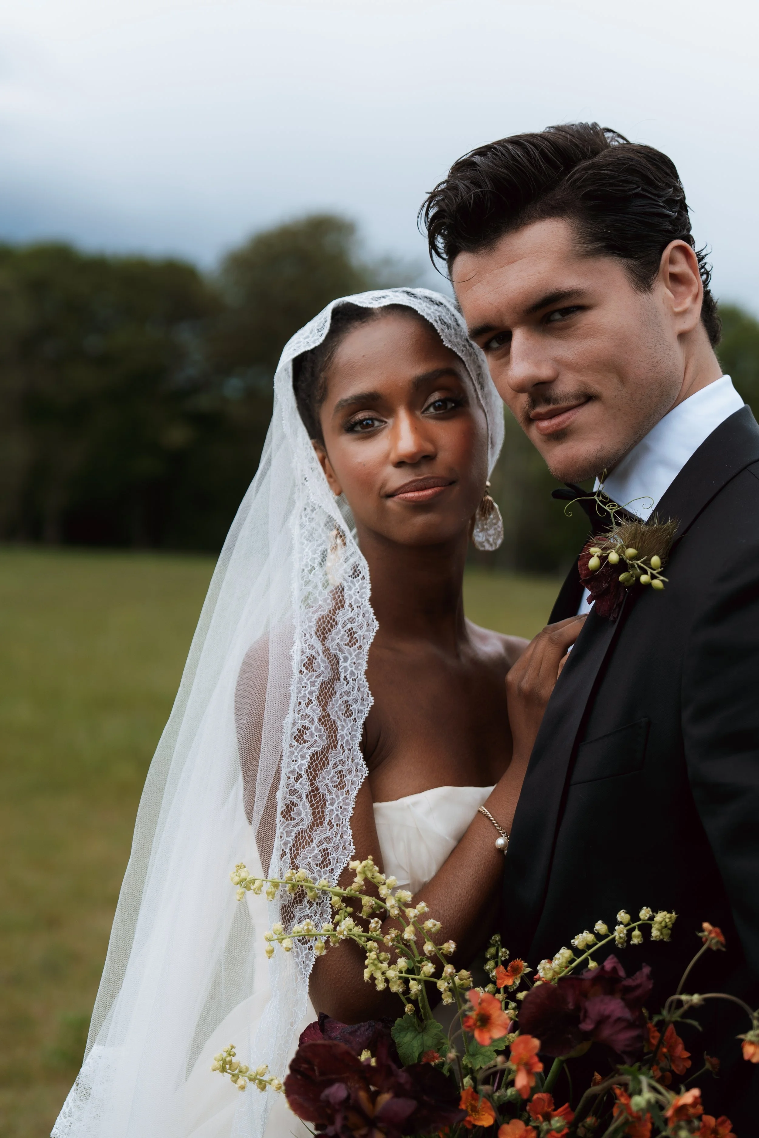 A bride and groom outdoors, close-up shot, the bride with lace wedding veil and Vera Wang gown, the groom in a tuxedo with bow tie, holding a colorful bouquet of flowers, greenery, and dark leaves. Background of trees and cloudy sky.