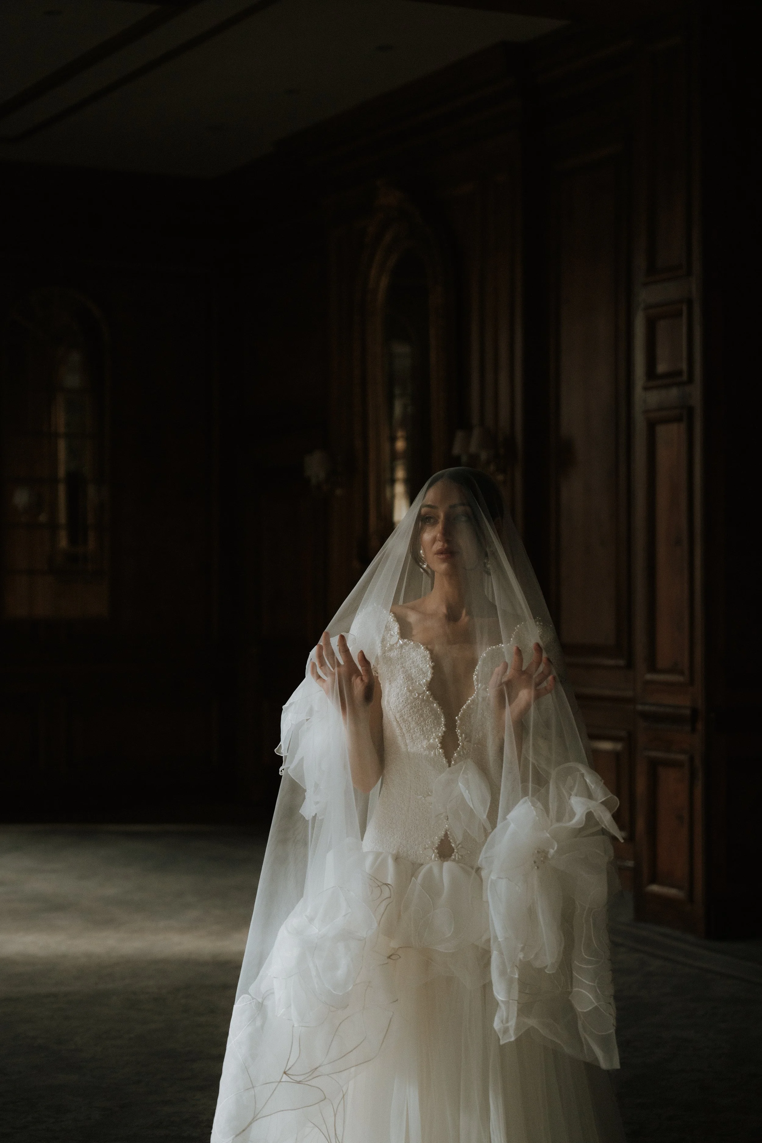 An Editorial bride in a white wedding dress and veil standing in a dark, ornate room with wooden walls and tall windows.