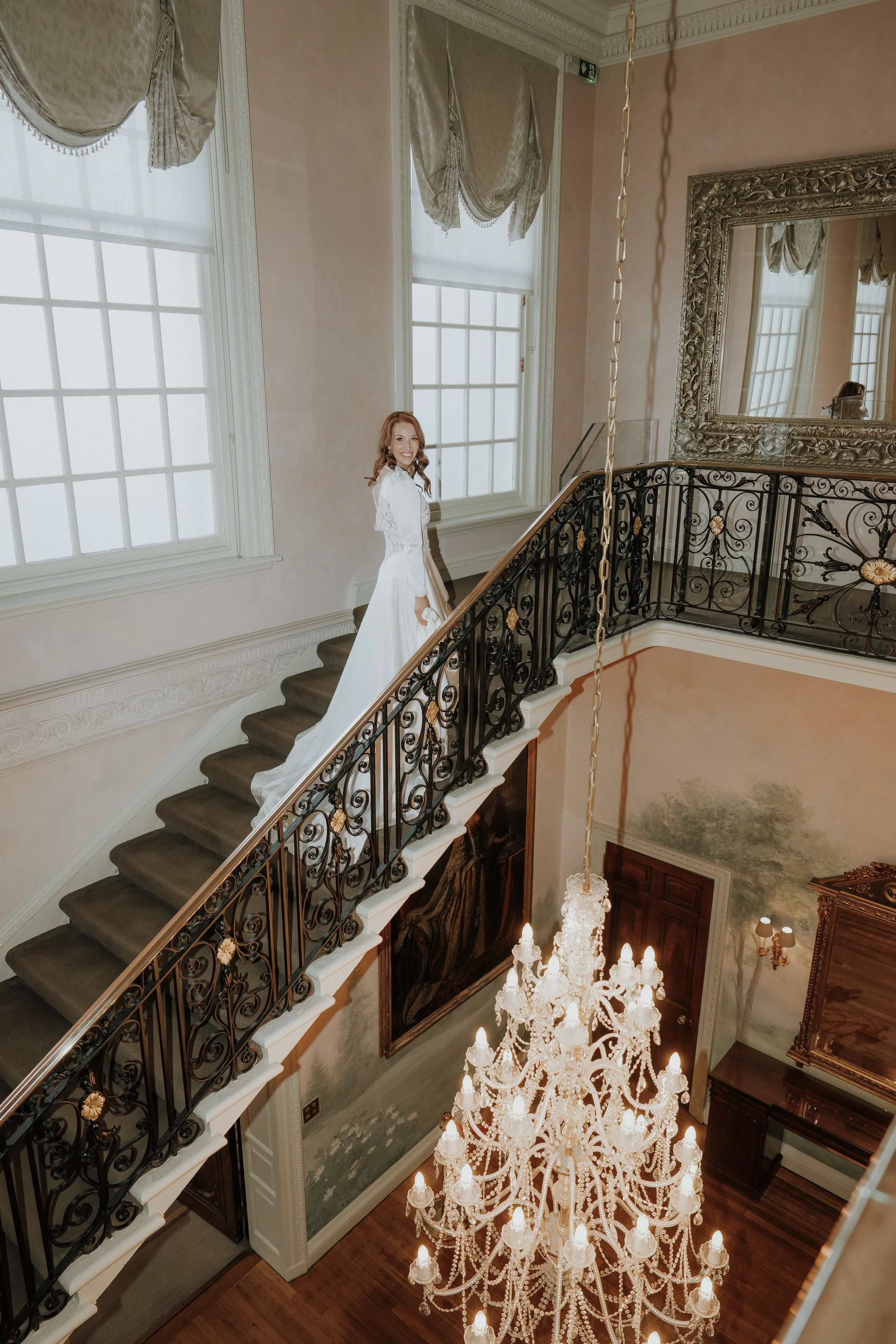 A bride in a white wedding dress standing on a staircase in an elegant, historic mansion with ornate iron railings and a grand chandelier.