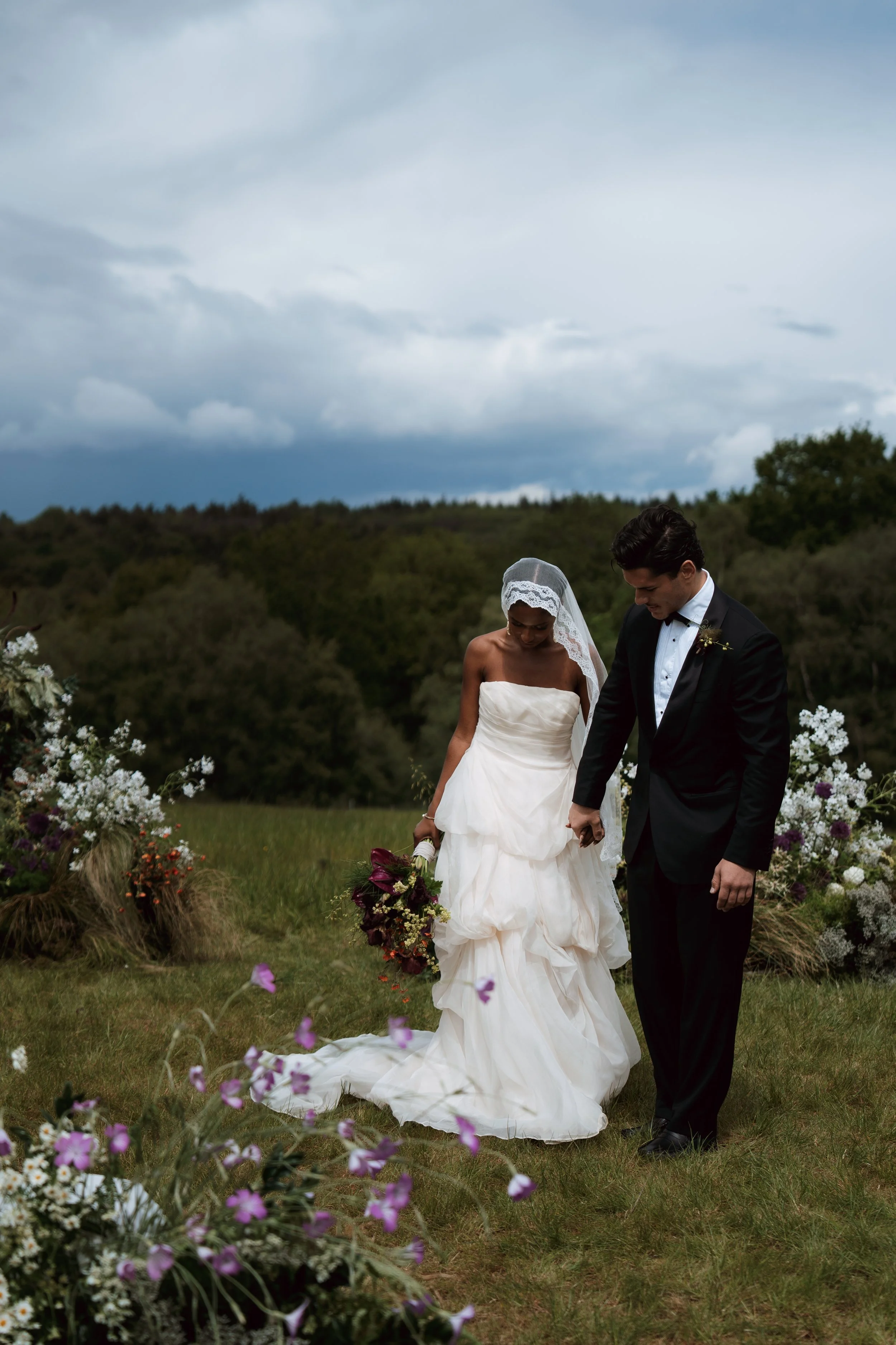 A bride and groom holding hands during an outdoor wedding ceremony, with floral arrangements and a cloudy sky in the background.