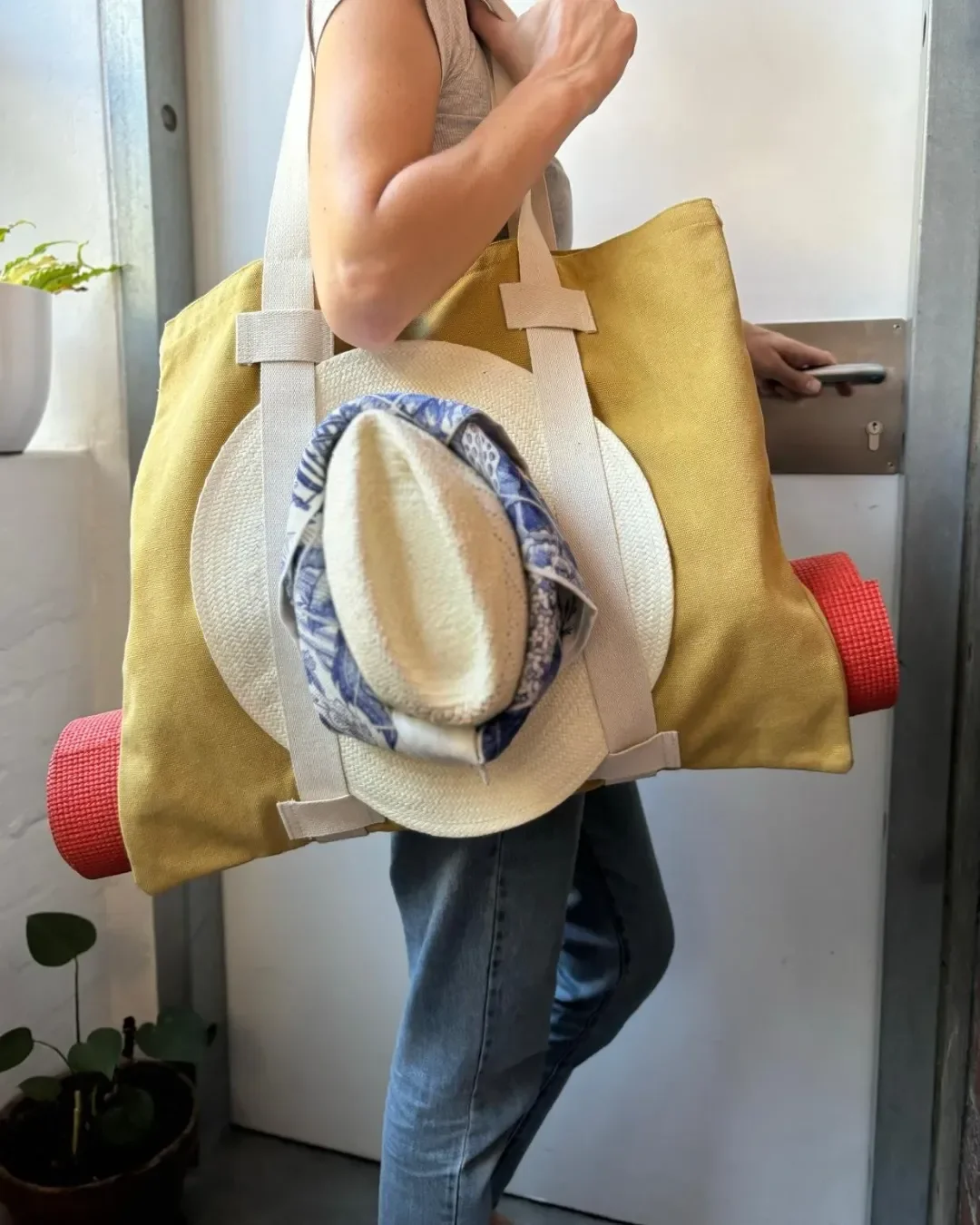 A woman carrying a mustard yellow Vienna Tote bag handmade in Cape Town by Soli for a custom yoga or pilates bag merchandise or corporate gift, featuring a yoga mat and sun hat secured by cotton straps. 