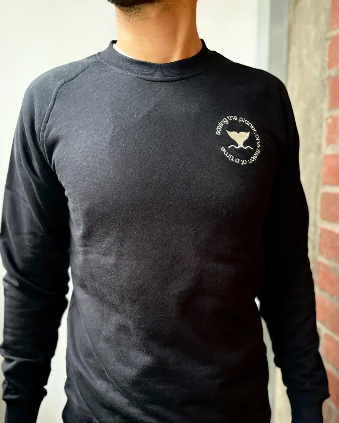 A man wearing a premium black 100% cotton sweater with a white embroidered circular whale tail logo, handmade by local artisans in Cape Town by Soli for corporate gifts & merchandise.