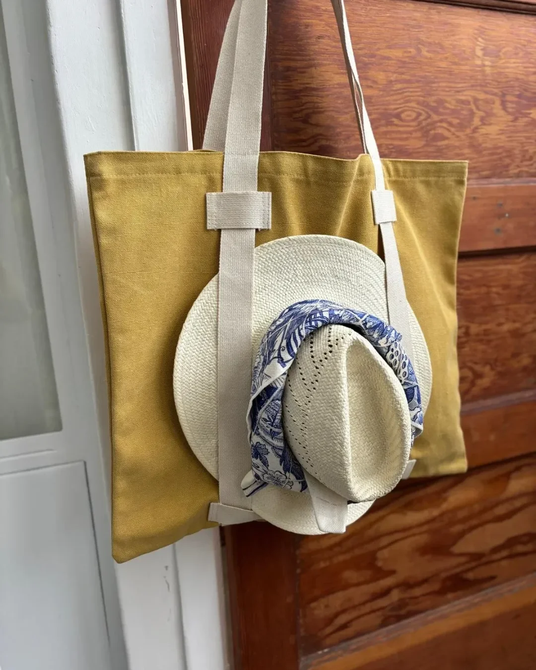Mustard yellow heavyweight canvas Vienna Tote hanging on a wooden door, featuring cotton straps securing a woven sun hat with a blue patterned scarf, handmade in Cape Town by Soli for a custom yoga or pilates bag merchandise or corporate gift.