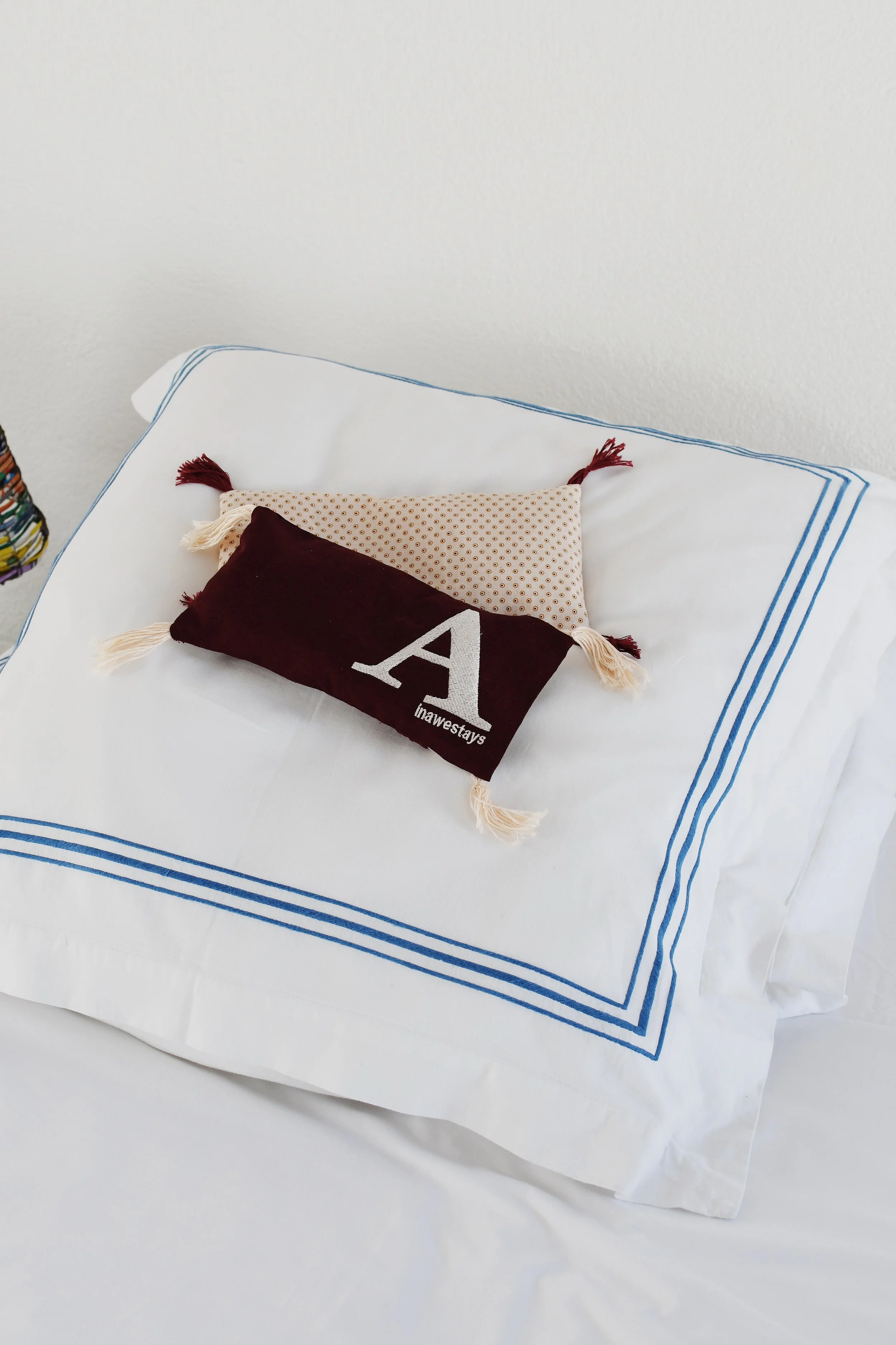Decorative pillows on a bed with white linens outlined in blue, featuring tassels and a letter 'A'.