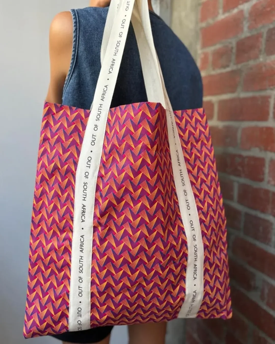 A person carrying a vibrant pink and purple geometric pattern 100% shweshwe tote bag, featuring a custom white cotton ribbon printed with 'OUT OF SOUTH AFRICA', handmade by local artisans for Soli in Cape Town.