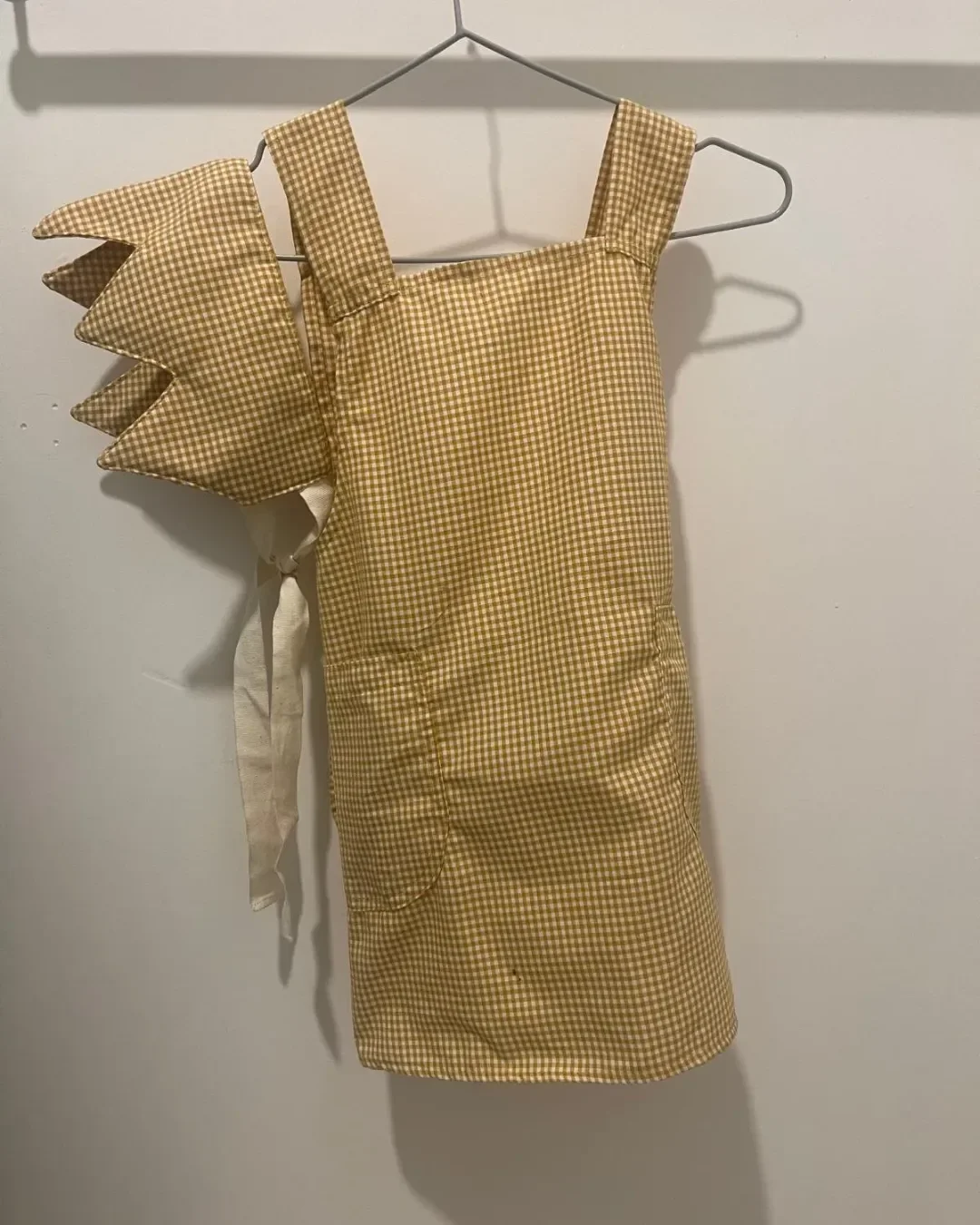 A matching set featuring an ochre gingham kids' apron and crown hanging on a silver hanger, representing sustainable hospitality amenities and custom corporate gifts from Soli, made in Cape Town.