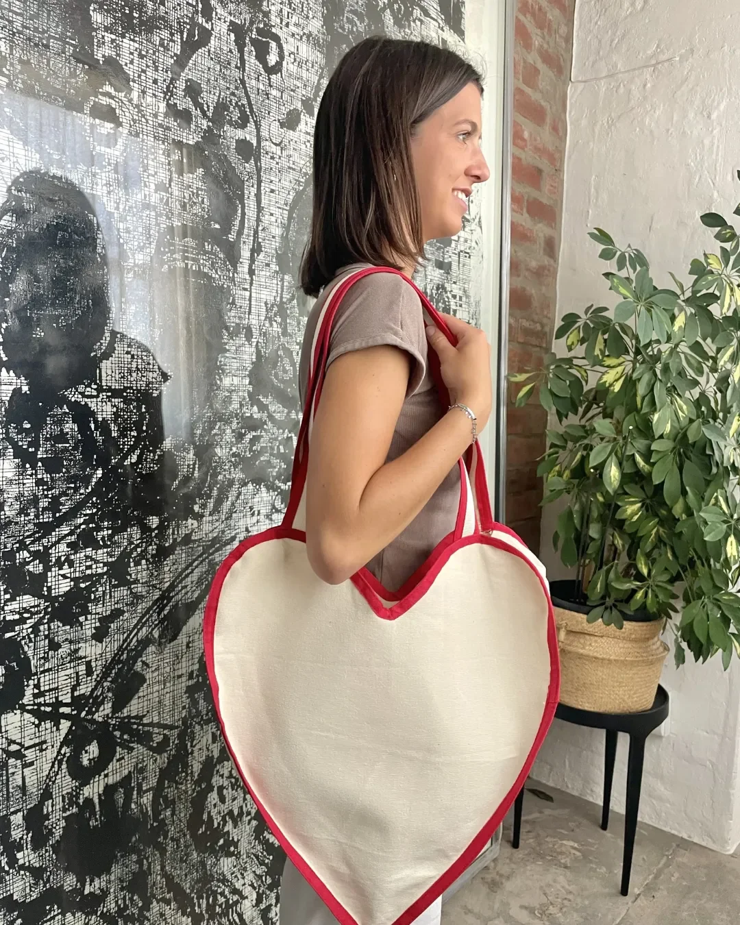A woman wearing a large cream heart-shaped canvas tote bag with red trim over her shoulder, handmade by Soli in Cape Town.