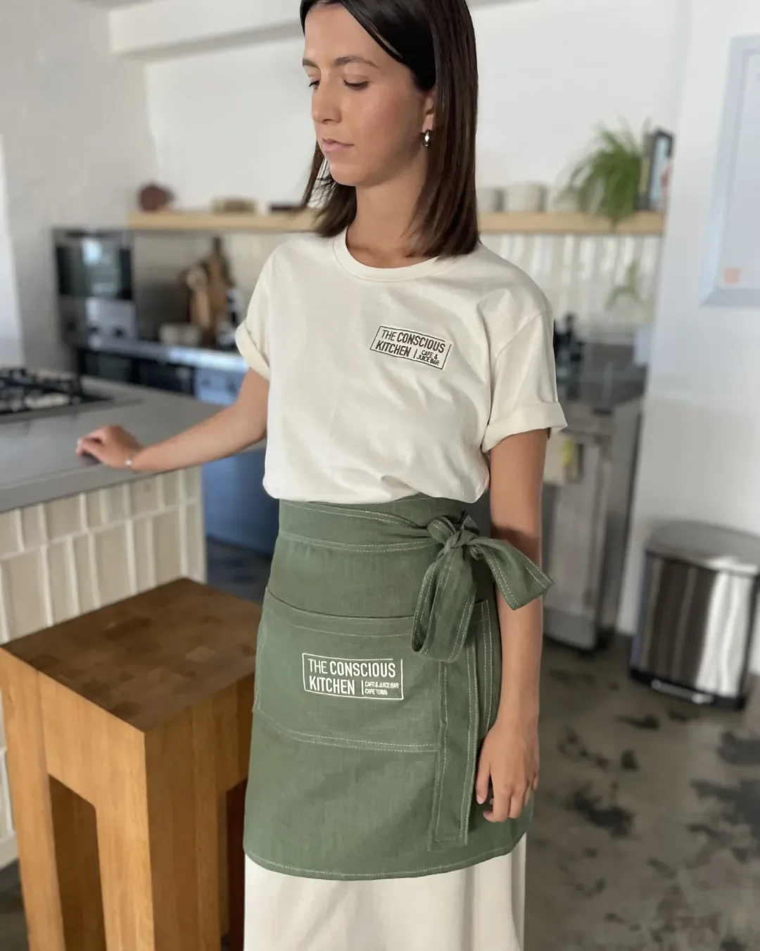 A woman wearing an olive green sustainable canvas half apron with "The Conscious Kitchen" embroidered logo, handmade by local artisans in Cape Town for hospitality use.
