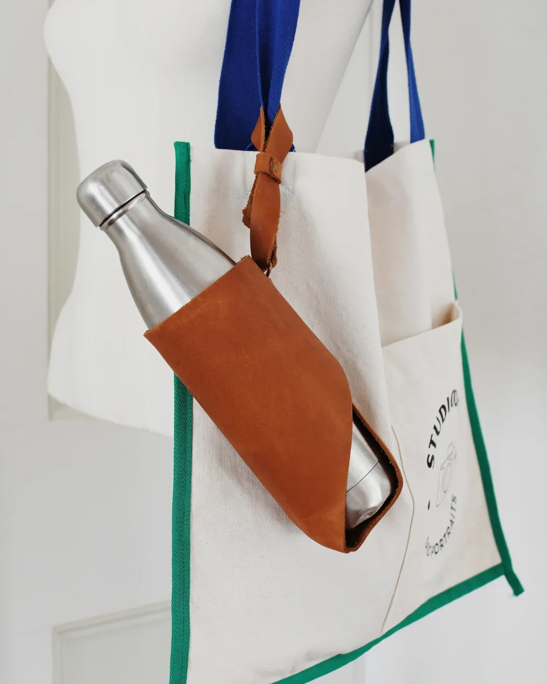 A high-quality tan leather water bottle holder attached to a canvas tote bag, featuring a sleek stainless steel bottle, Handmade by Soli in Cape Town for sustainable merchandise or corporate gifts.