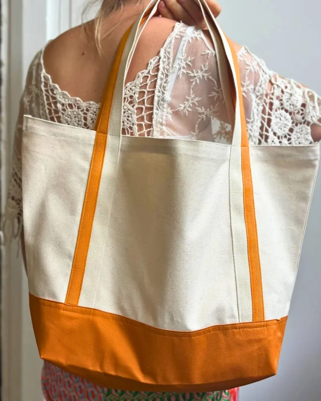 Woman wearing a white lace top carrying a large cream canvas Milano tote bag over her shoulder, featuring a bright orange bottom panel and matching orange accents on the straps.