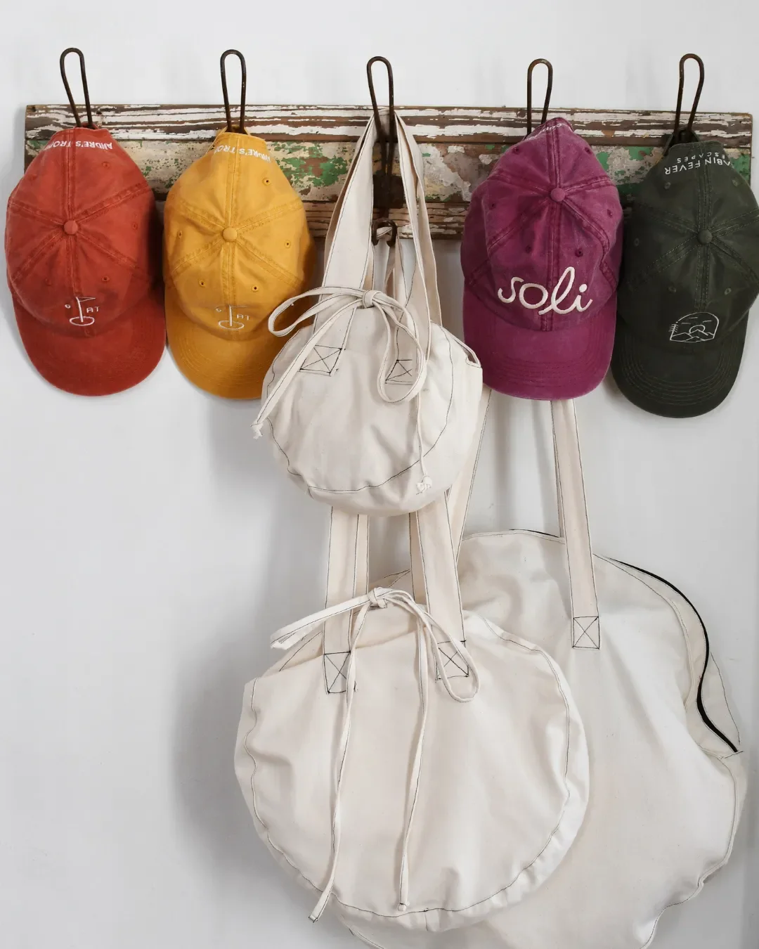 A collection of small, medium, and large London circular canvas bags hanging on hooks alongside colorful branded caps, Handmade by Soli in Cape Town for sustainable merchandise or corporate gifts.