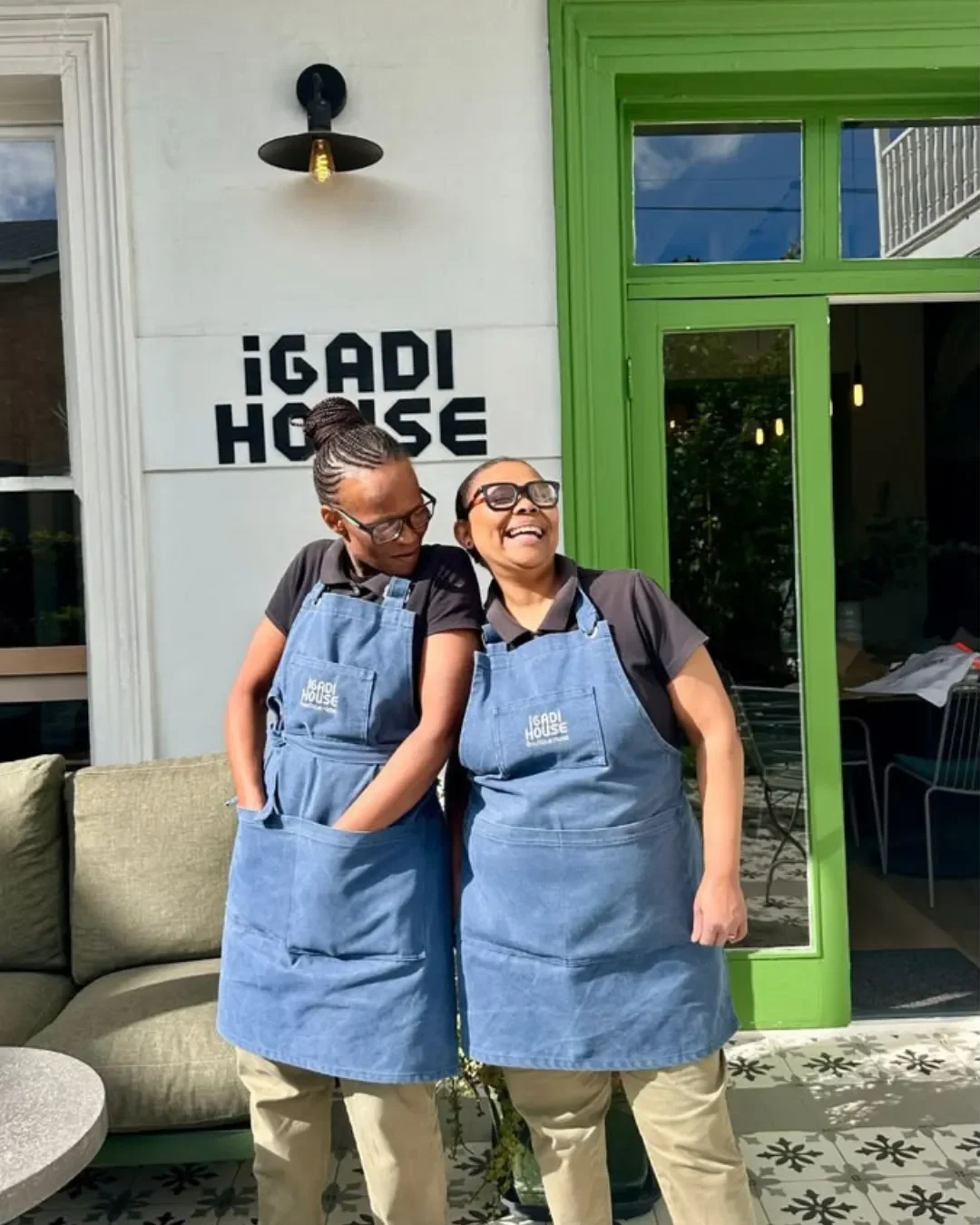 Two staff members at iGadi House are wearing matching custom-branded blue canvas hospitality aprons with embroidered logos, handmade in Cape Town by Soli.
