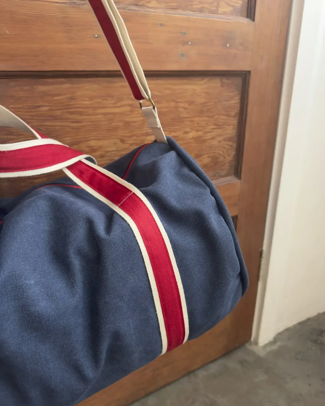 A navy blue canvas duffel bag with cream and red striped shoulder straps hanging against a wooden door, handmade by Soli in Cape Town for sustainable retail merchandise.