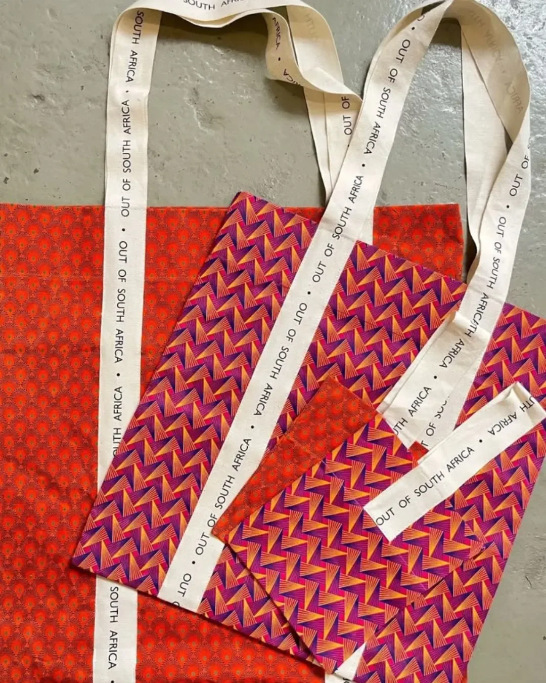 A flat lay of two vibrant 100% shweshwe tote bags in orange and purple geometric patterns, featuring long custom white cotton ribbons printed with 'OUT OF SOUTH AFRICA', handmade by local artisans for Soli in Cape Town.
