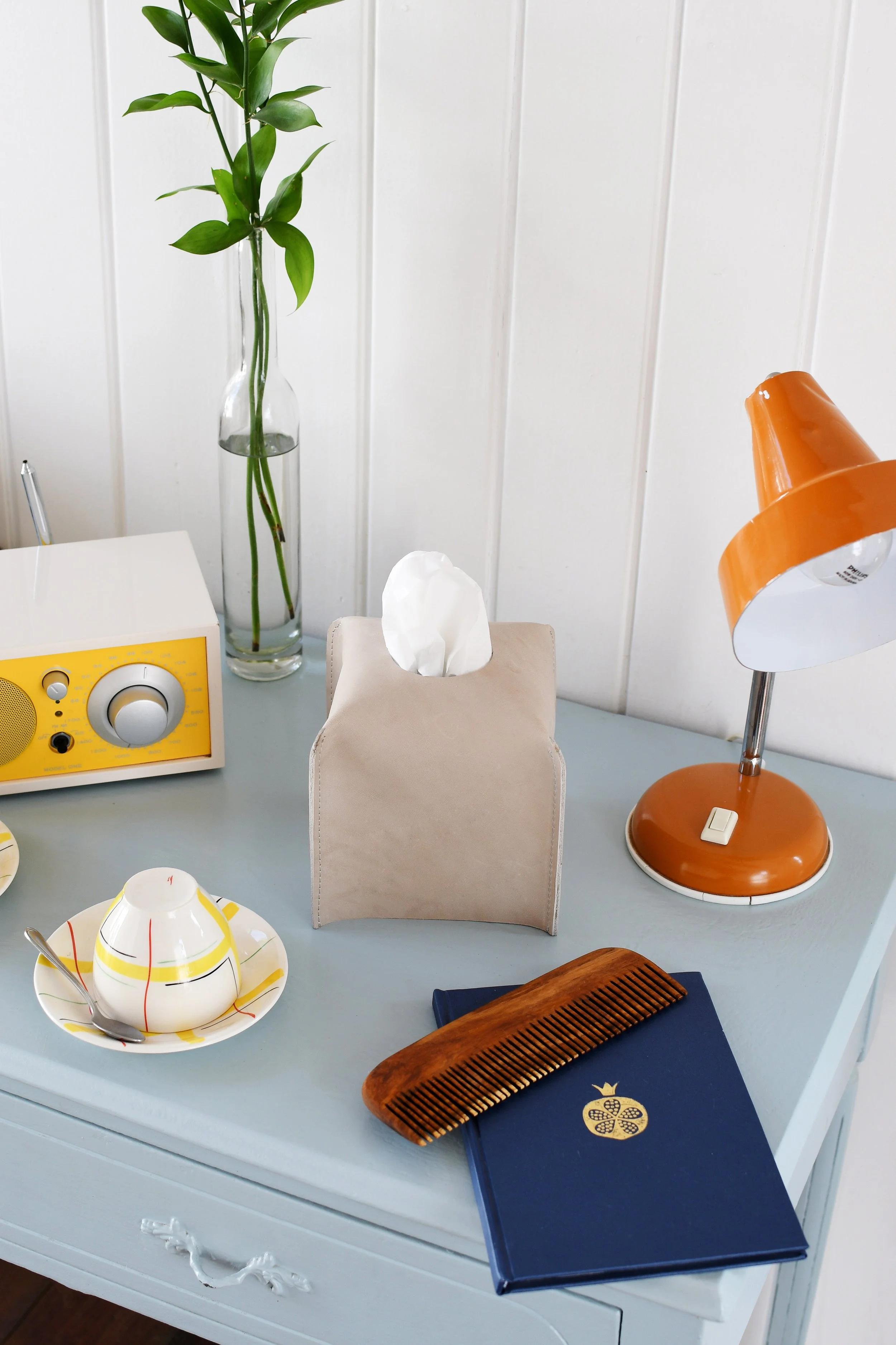 Desktop with a radio, tissue box, vintage lamp, notebook, wooden comb, teacup, saucer, and a vase with leaves.